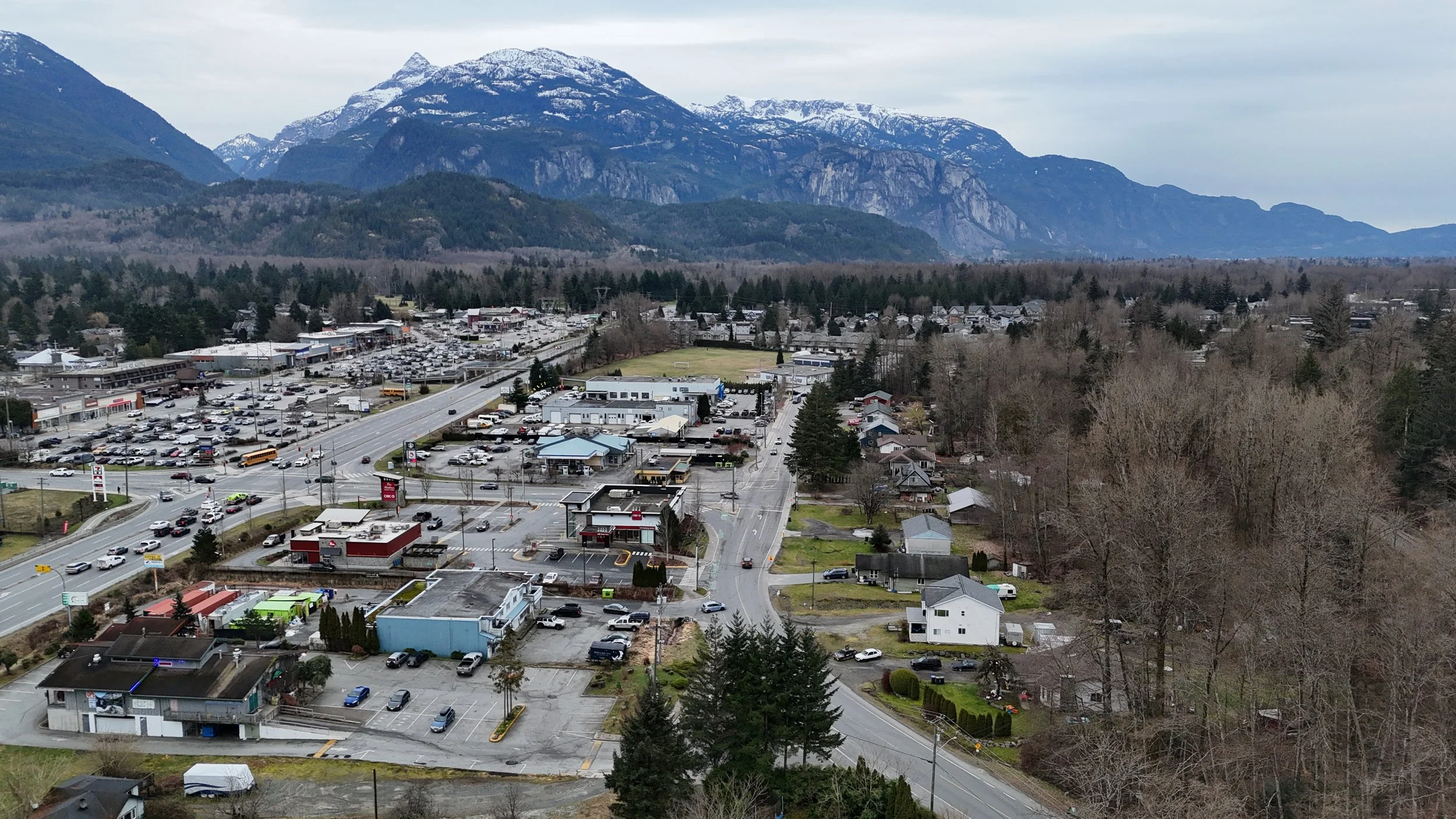 Aerial view of a small town with shops, residential houses, parking lots, and trees, with snow-capped mountains in the background.