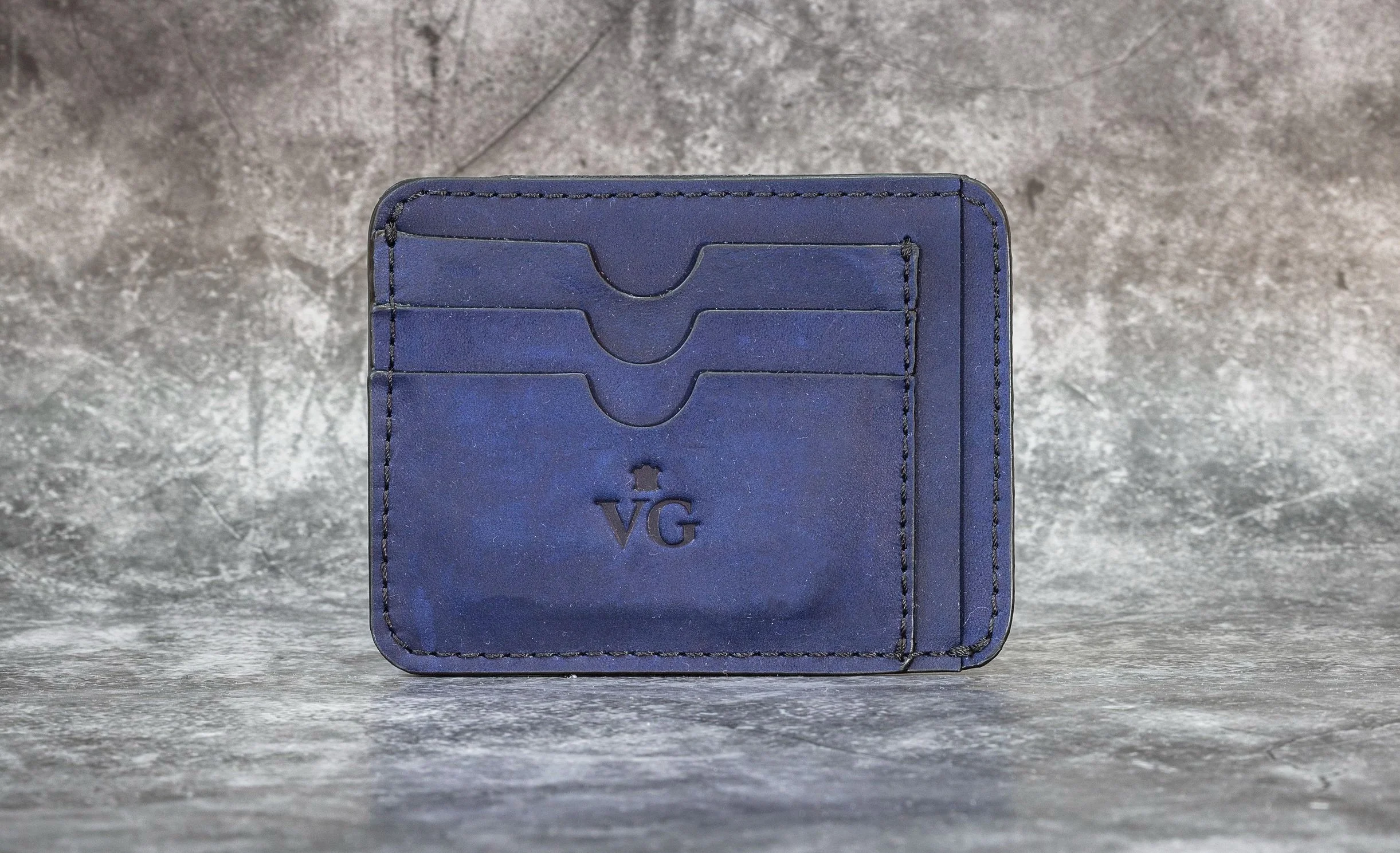 A blue leather wallet with three card slots and the initials 'VG' embossed on the front, placed on a textured gray surface.