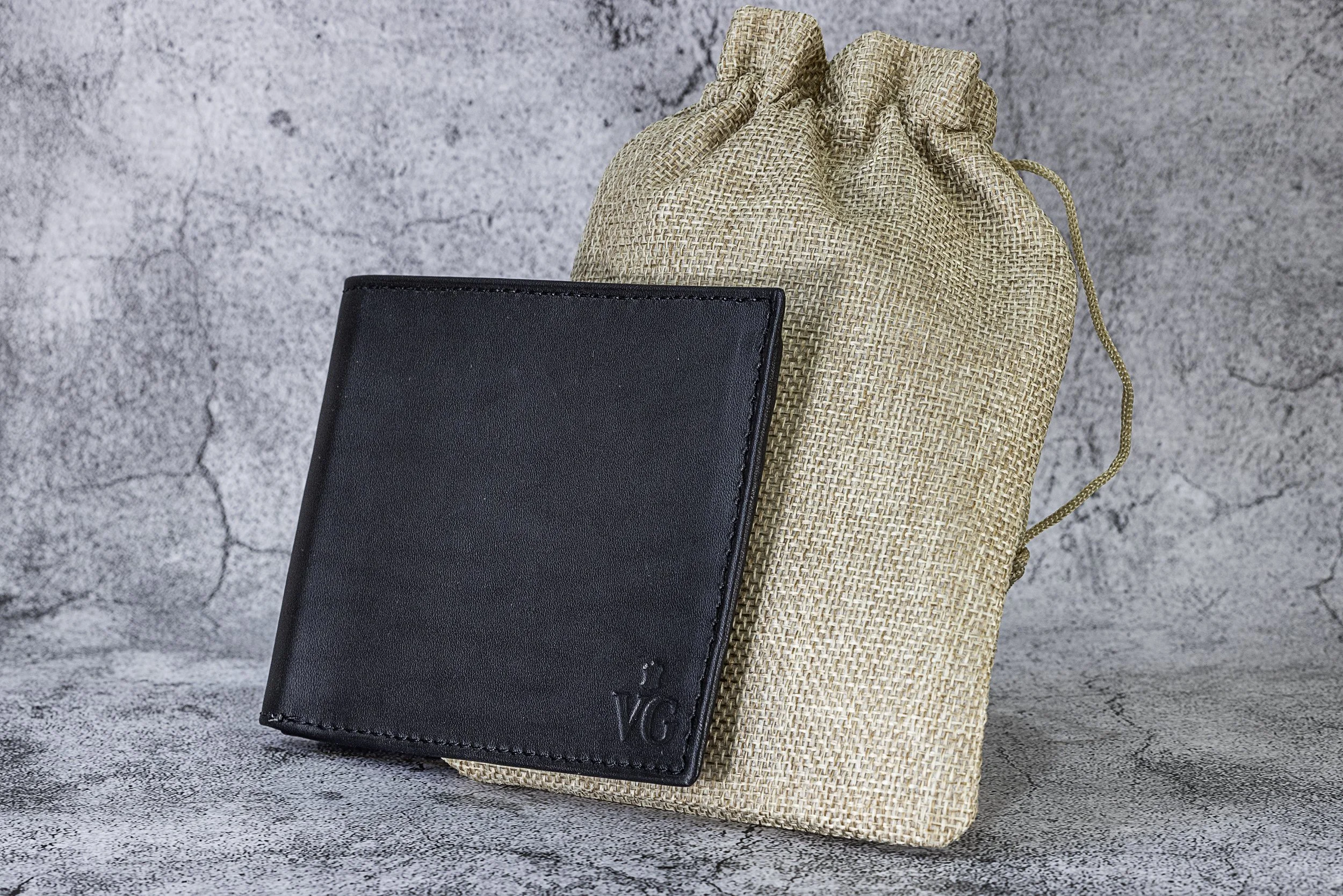 Black leather wallet with embossed lettering next to a beige woven pouch, placed on a textured gray surface.