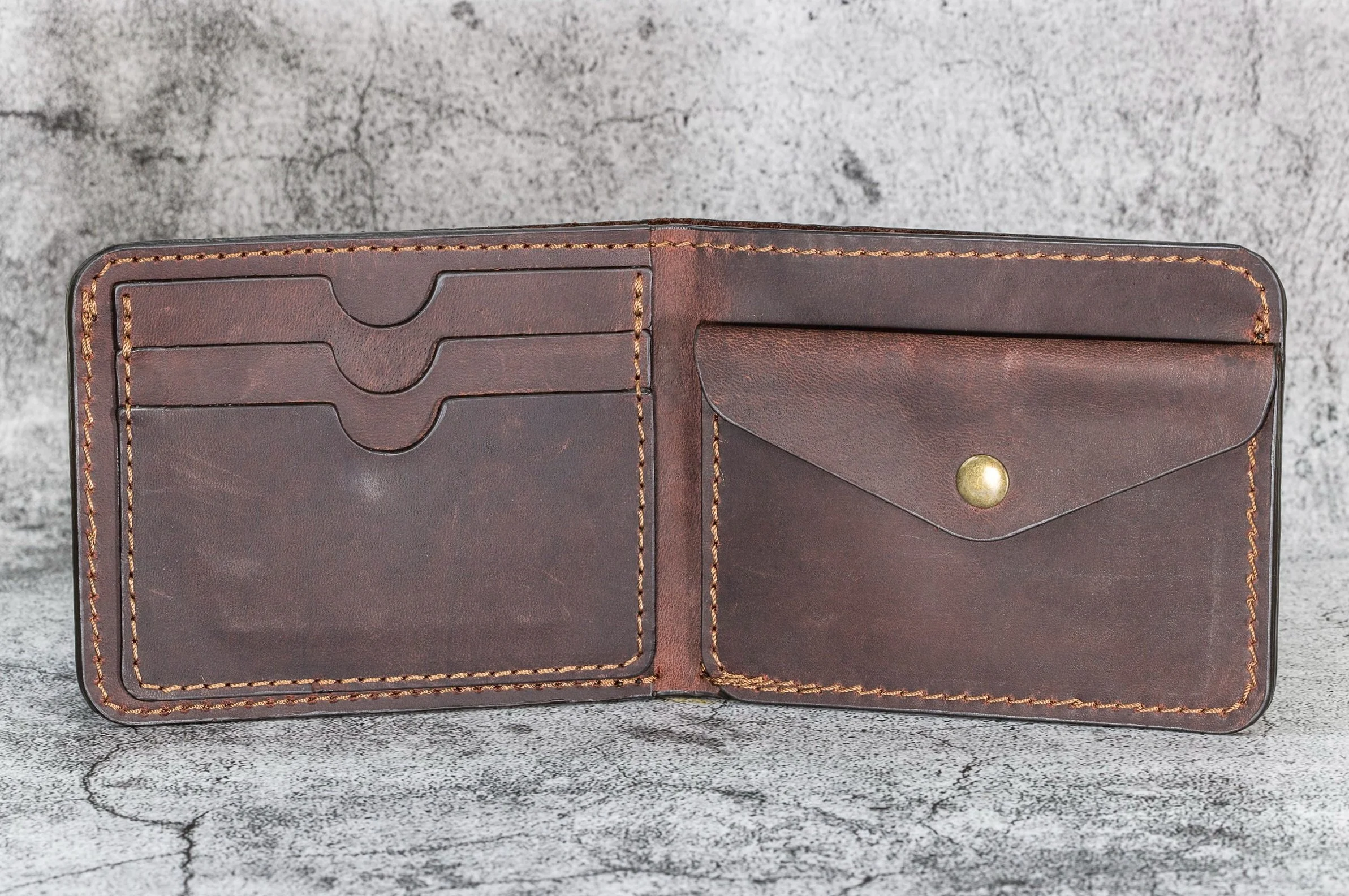 Open brown leather wallet with card slots and a snap button pocket, placed on concrete surface.