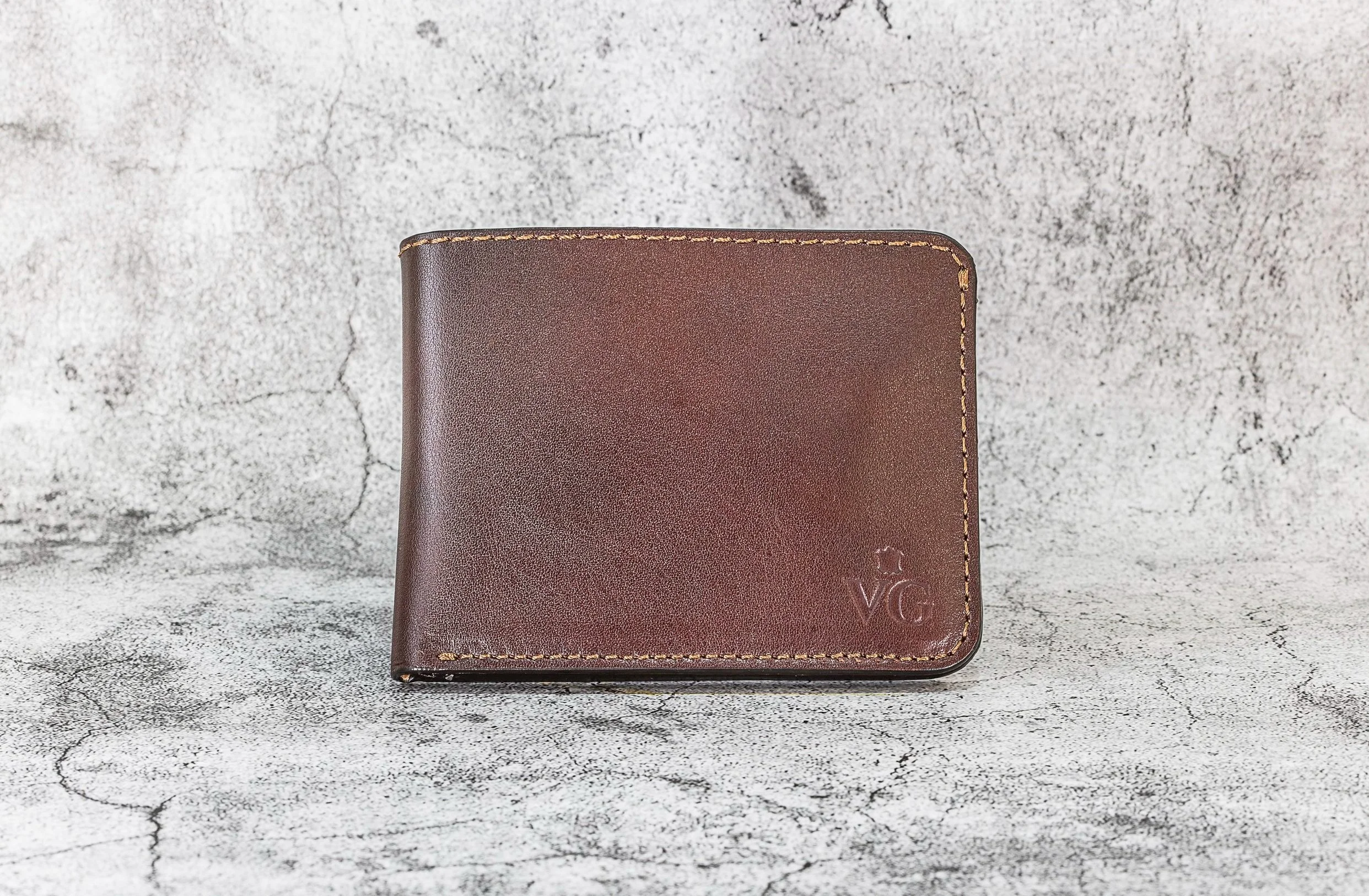 Brown leather wallet with V G embossed on front, placed on a textured gray surface.