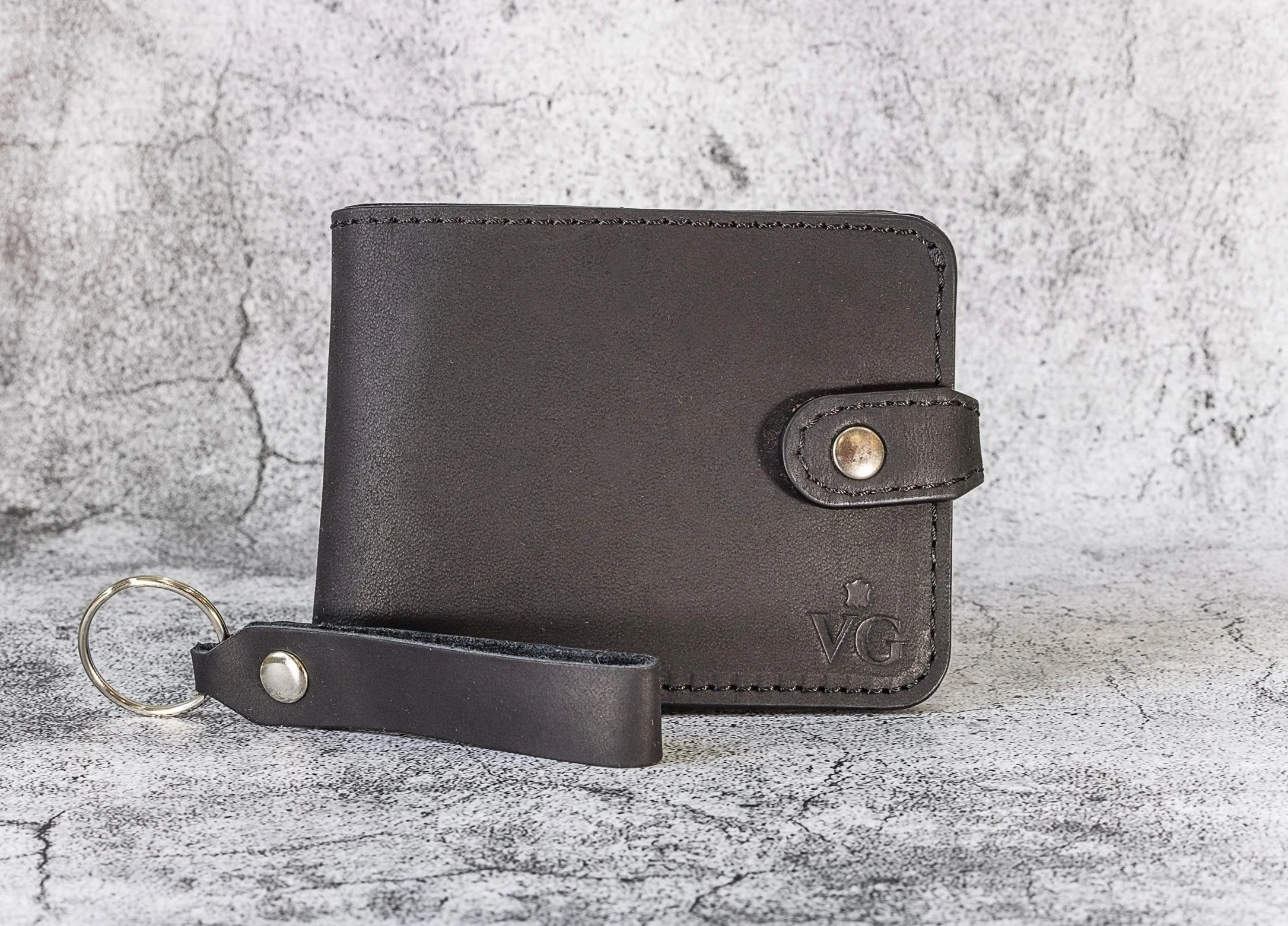 Black leather wallet with a snap closure and a leather keychain attached, embossed with the initials 'VG', set against a concrete background.