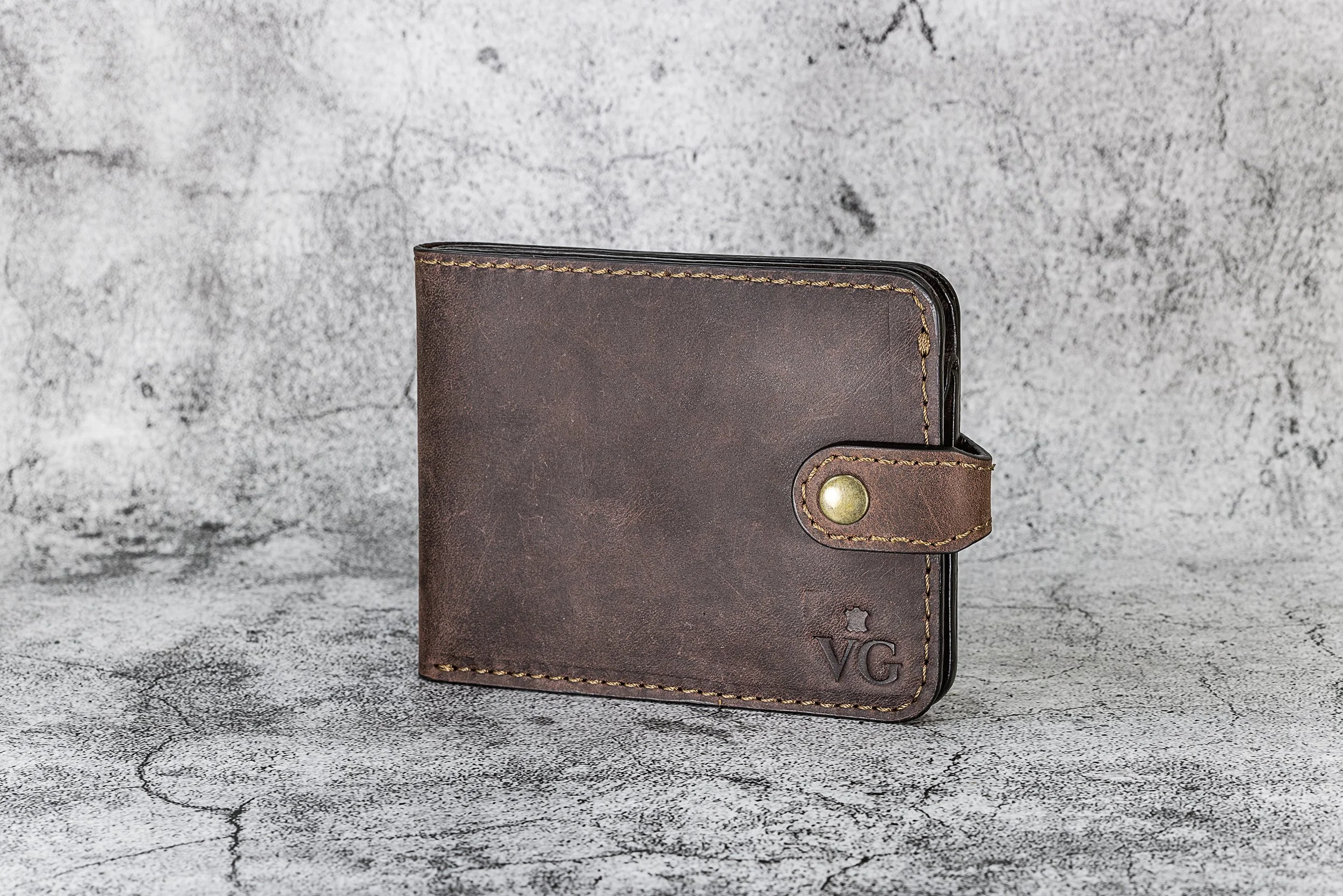 Brown leather wallet with a snap closure and gold-colored button, placed on a textured gray surface and background.