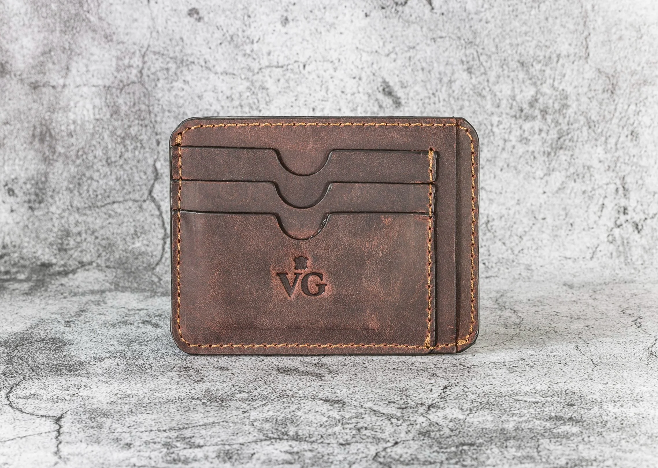 Brown leather wallet with card slots and initials 'VG' engraved on the front, placed on a gray textured surface.