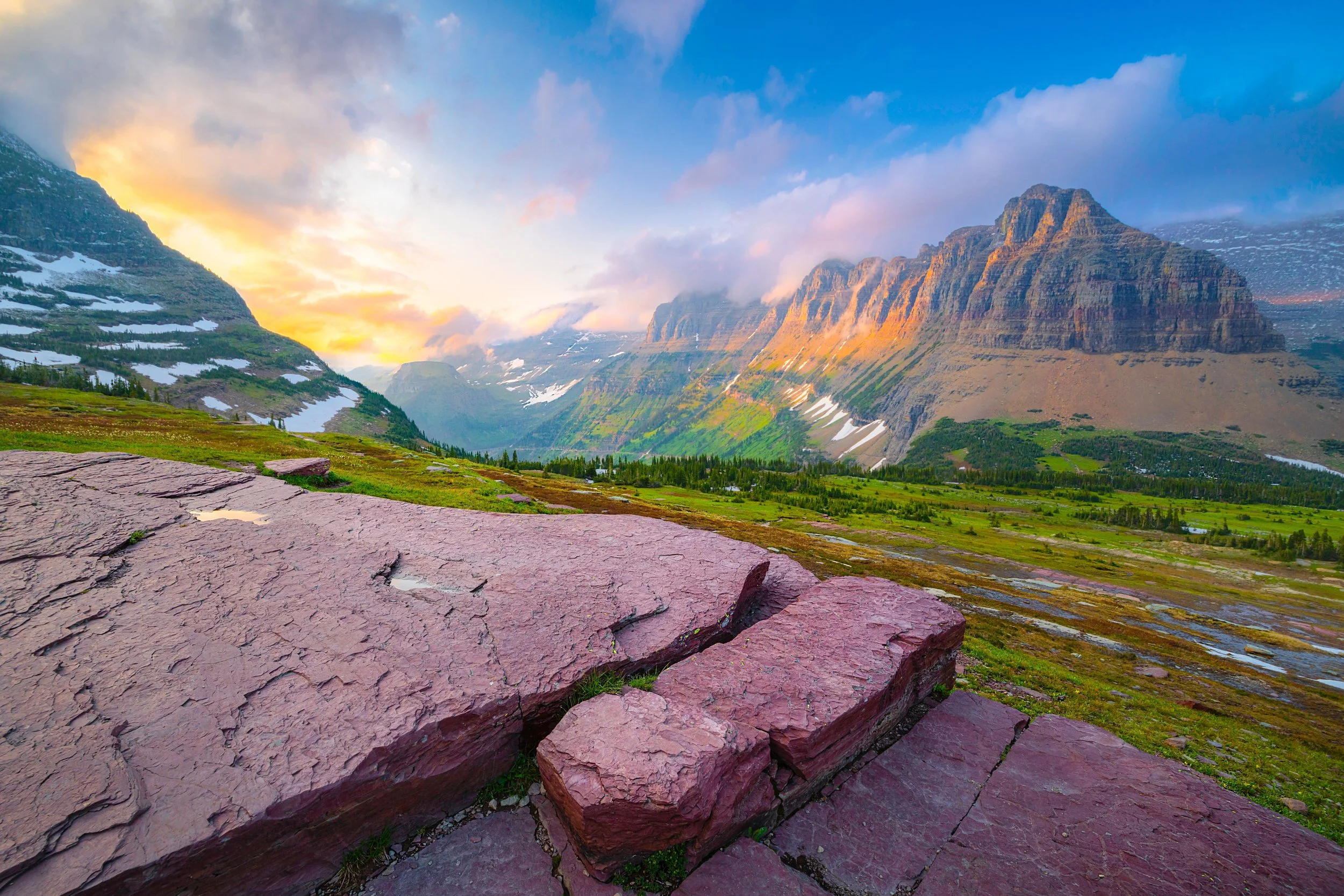 A scenic Montana landscape of mountains with snow patches, a green valley, and a colorful sunset sky.