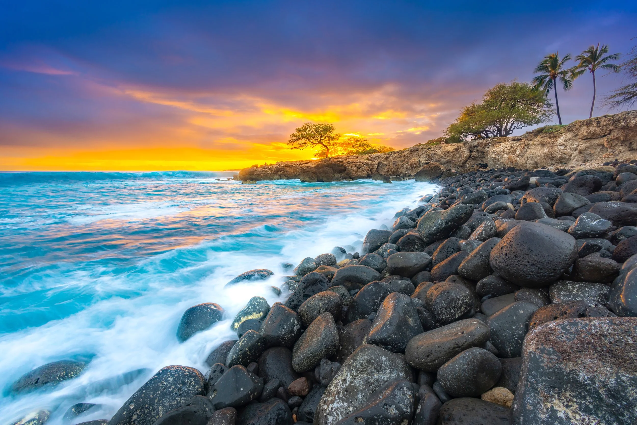 Sunset over a rocky Hawaiian coastline with waves crashing on black stones and trees on a cliff in the distance.