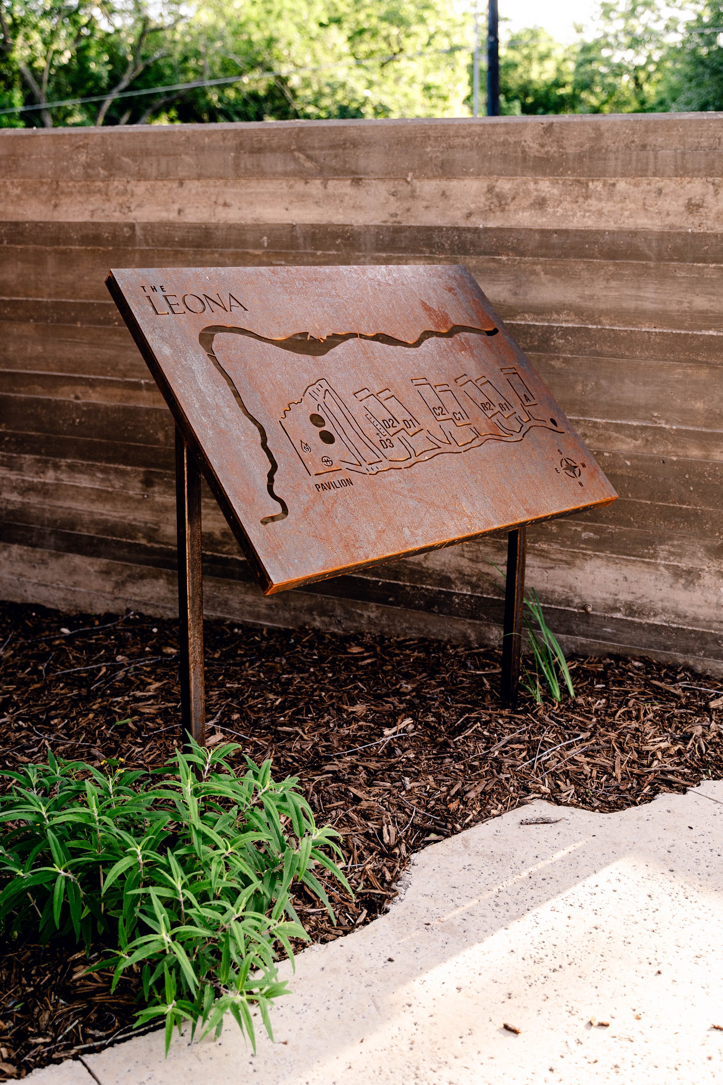 Rustic metal sign with the name "The Leona," displaying a map, surrounded by a concrete wall, mulch, and green plants.