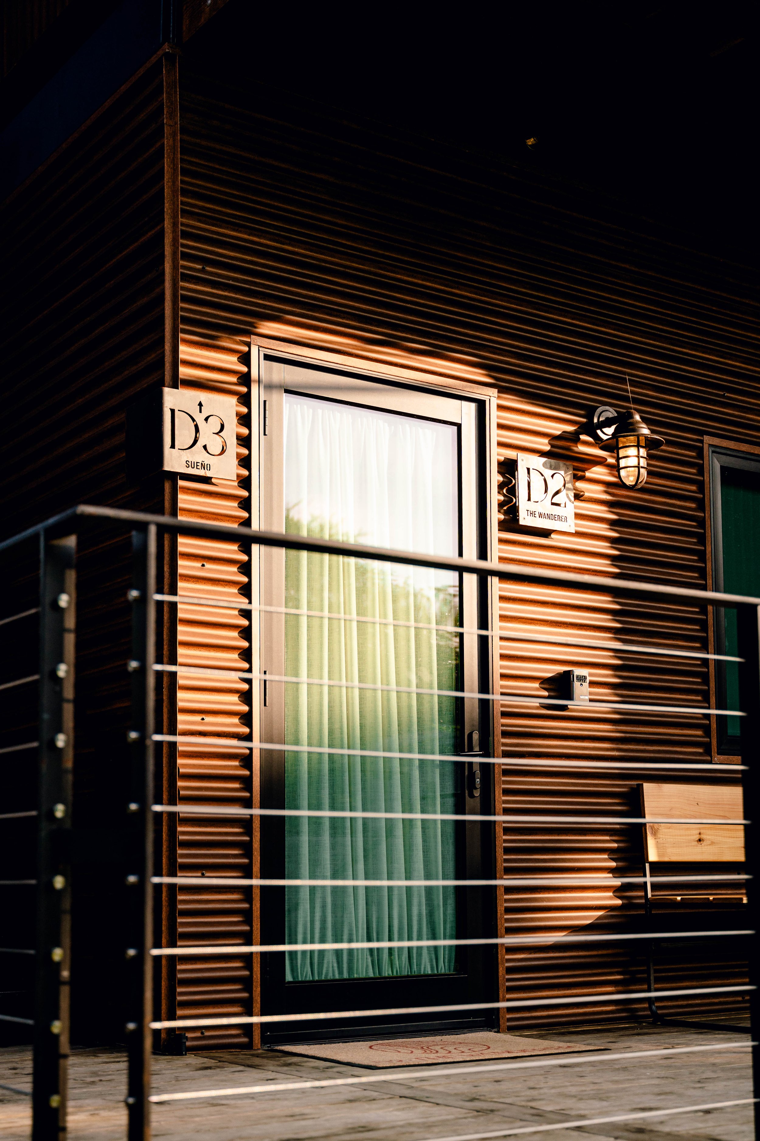 Modern building with corrugated metal siding, featuring a door labeled 'D3 Sueño' and 'D2 The Wanderer'; includes a railing and a bench.
