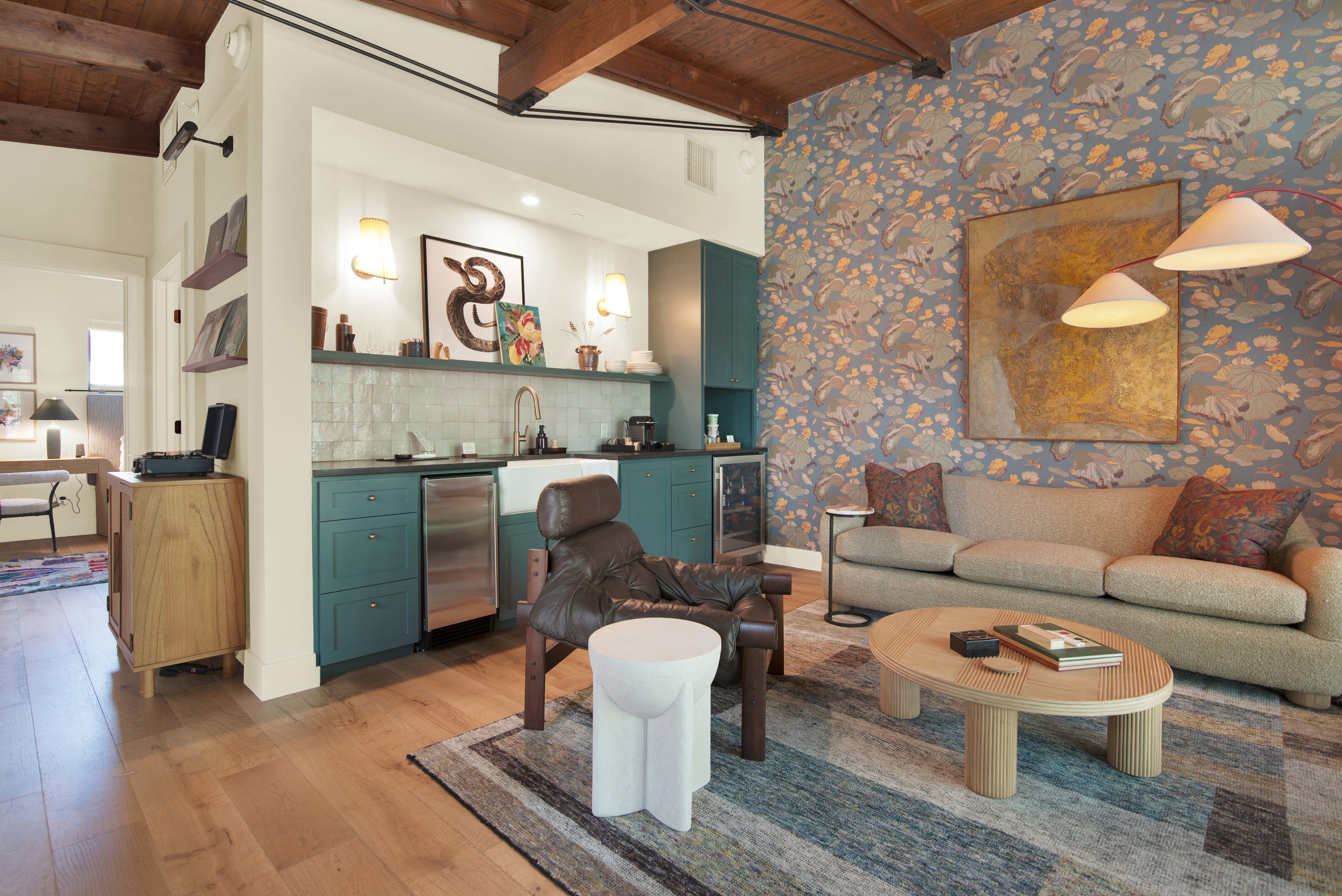 Cozy living room with a mix of modern and traditional decor. Features a teal cabinet kitchenette, patterned wallpaper, beige sofa, leather armchair, wooden coffee table, small white side table, and wall art. Wooden floors and exposed wooden beams com