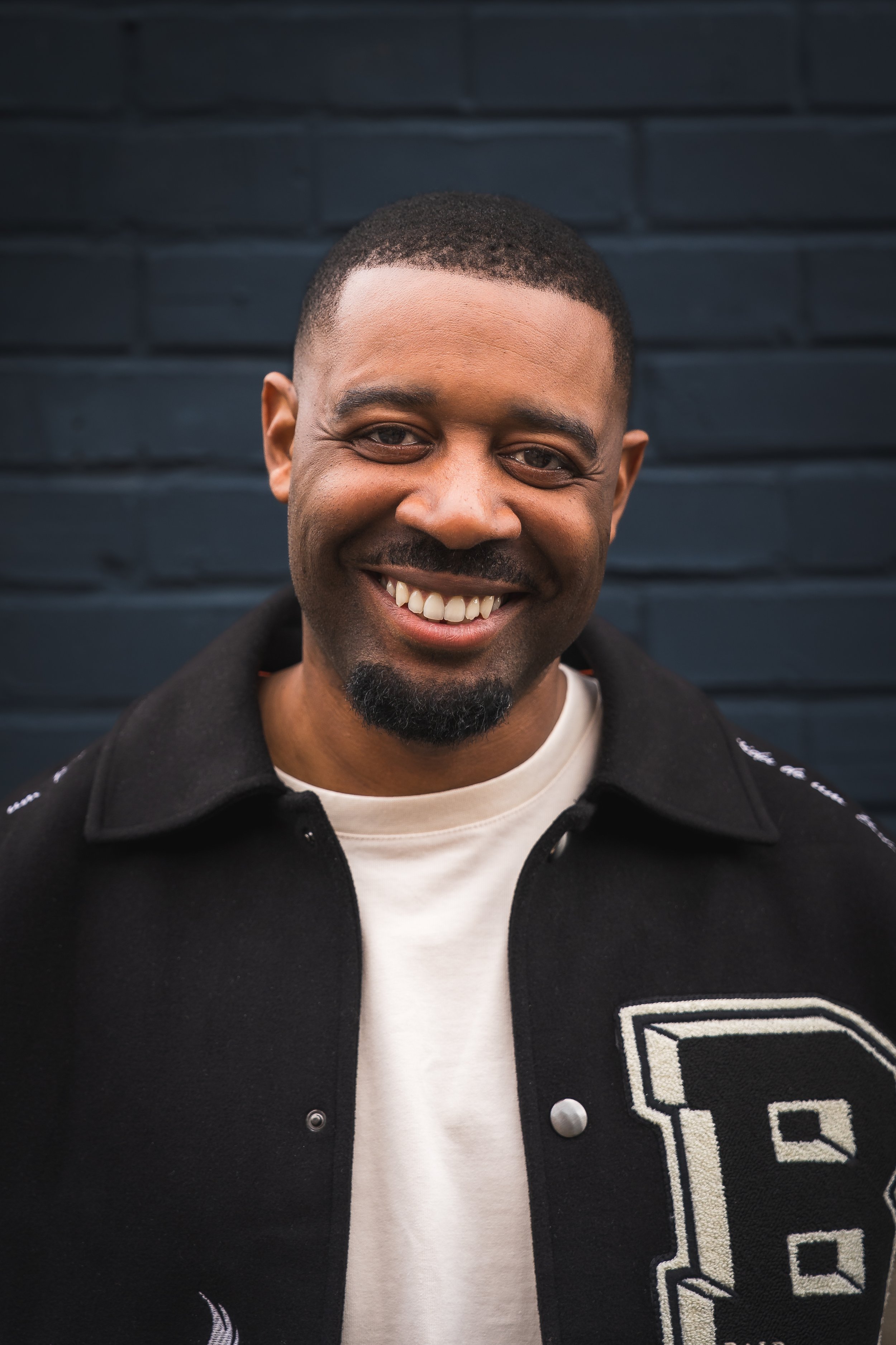 Close-up of a smiling man with short hair and a goatee, wearing a black jacket over a white shirt, standing in front of a dark brick wall.