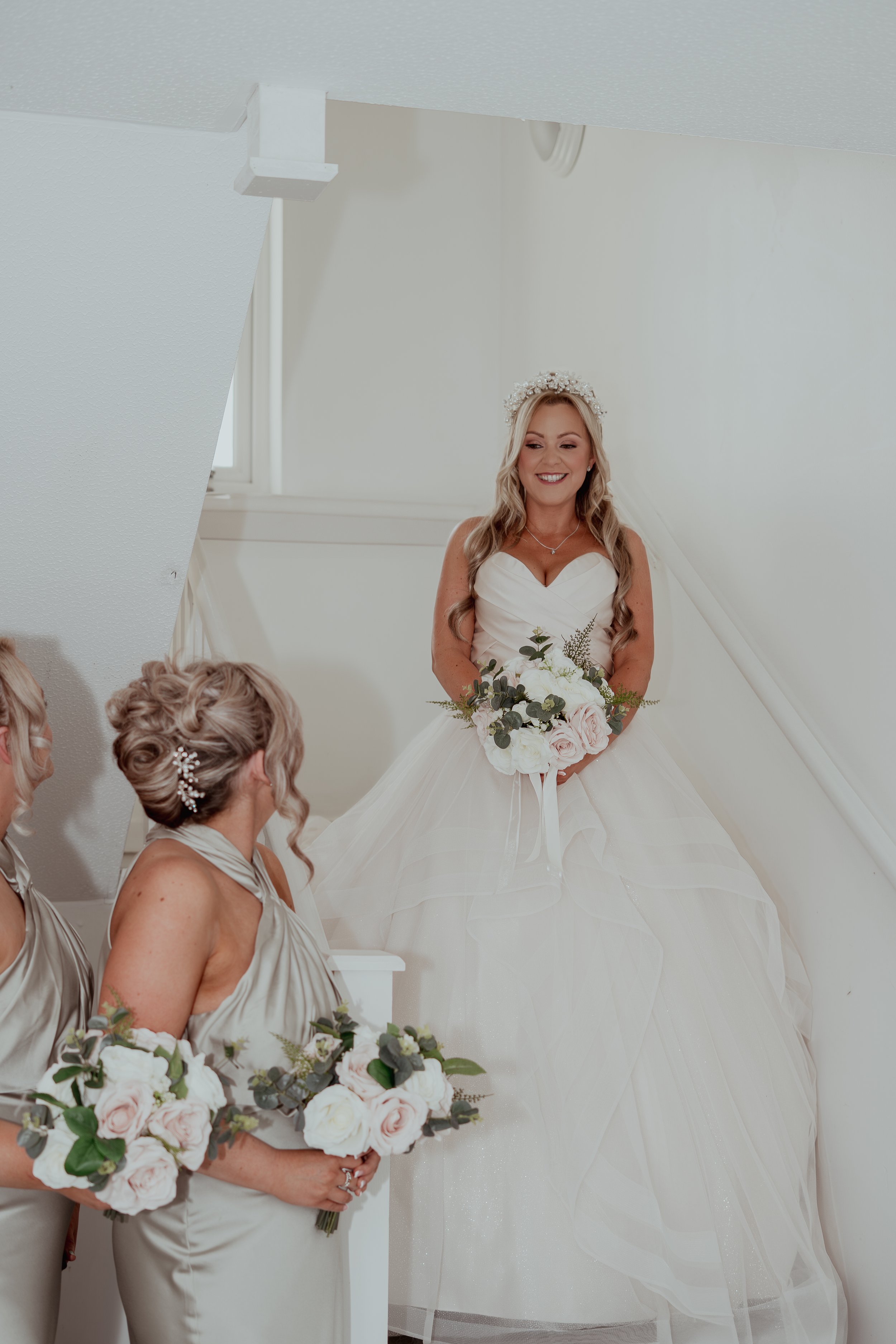 Bride in a white wedding gown holding a bouquet of pink and white roses, smiling in a stairwell, with bridesmaids in taupe dresses holding matching bouquets nearby.
