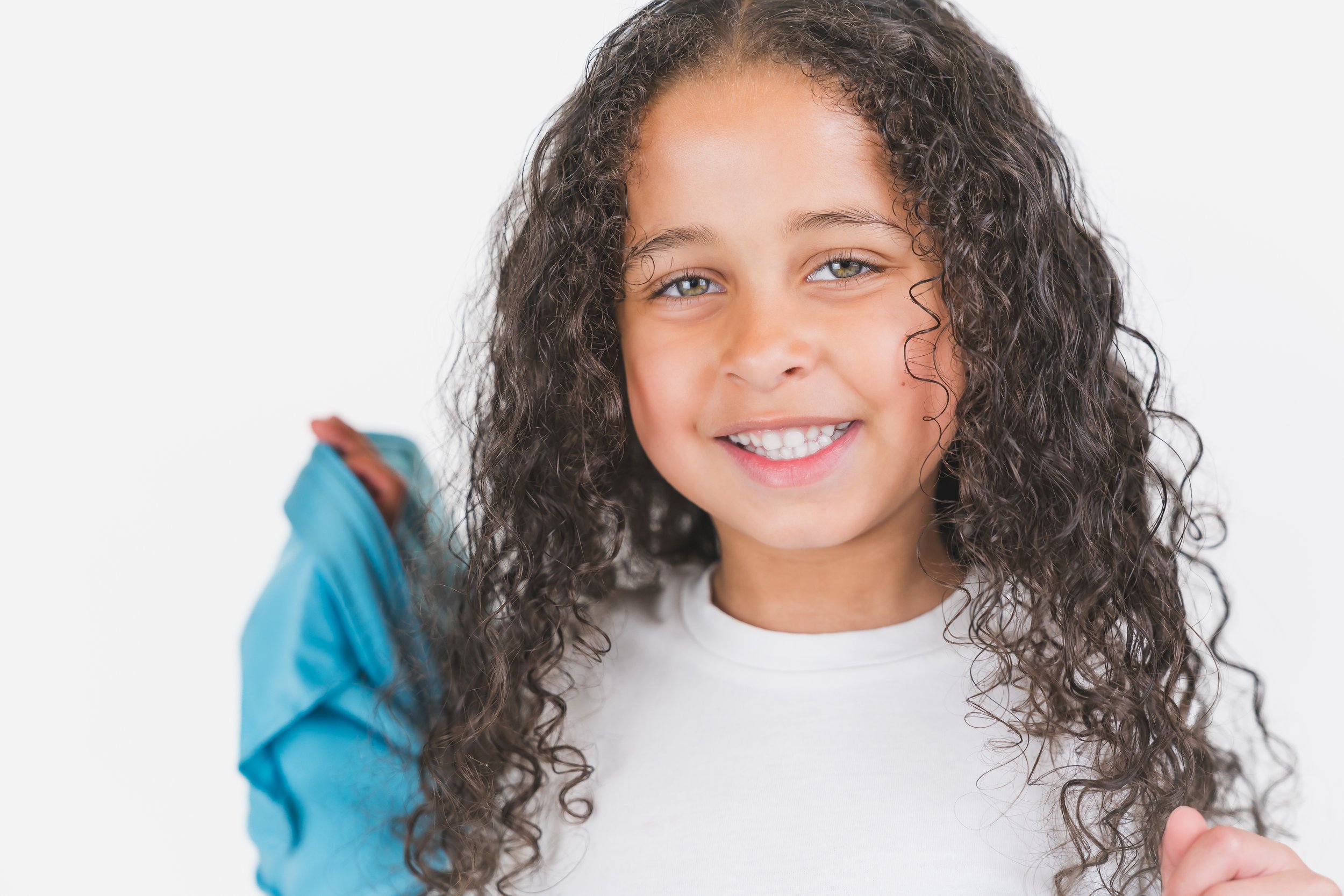 Young girl with long curly hair smiling and holding a blue backpack over her shoulder.
