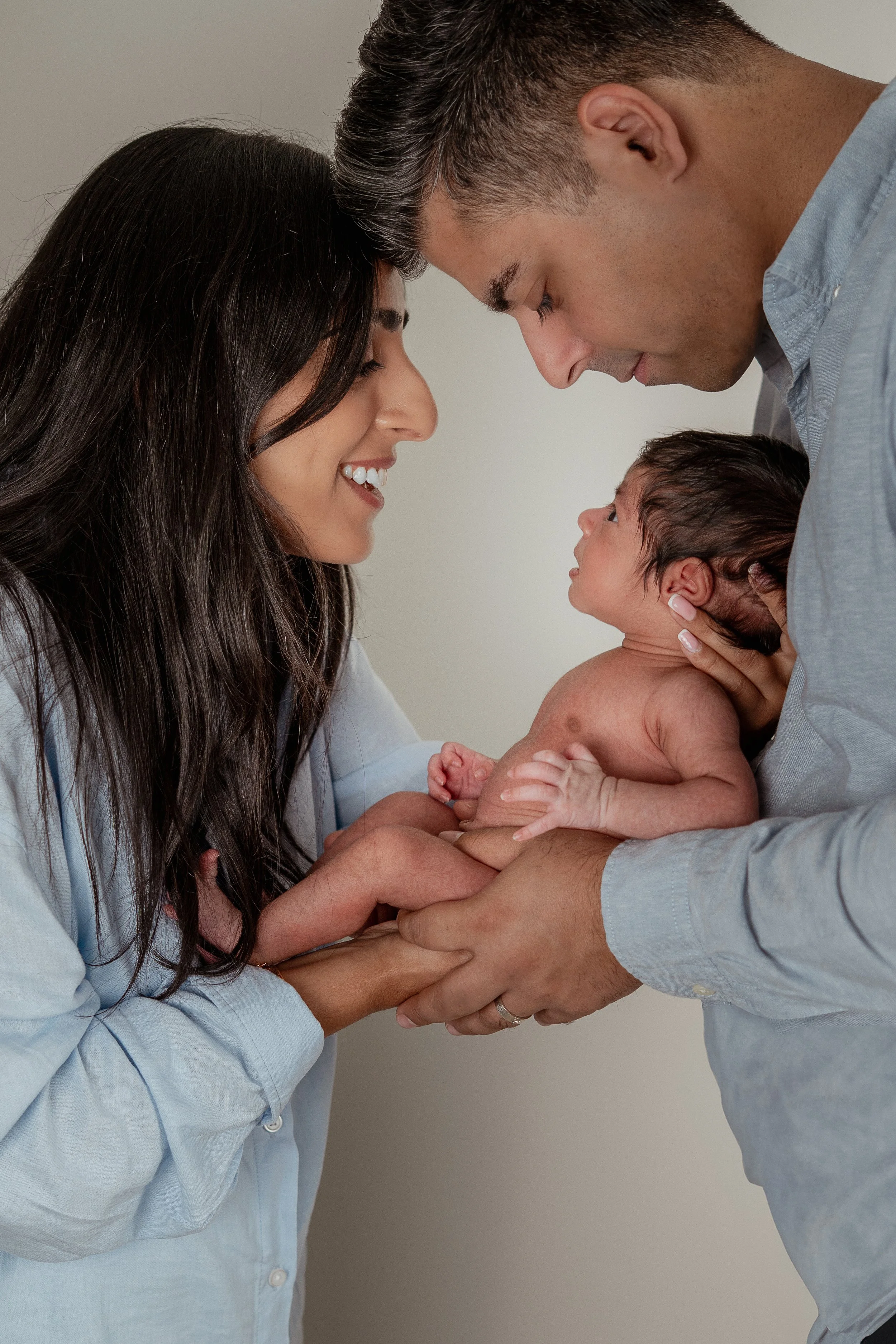 Photo of a family, dad holding baby against his chest whilst mum looks towards him