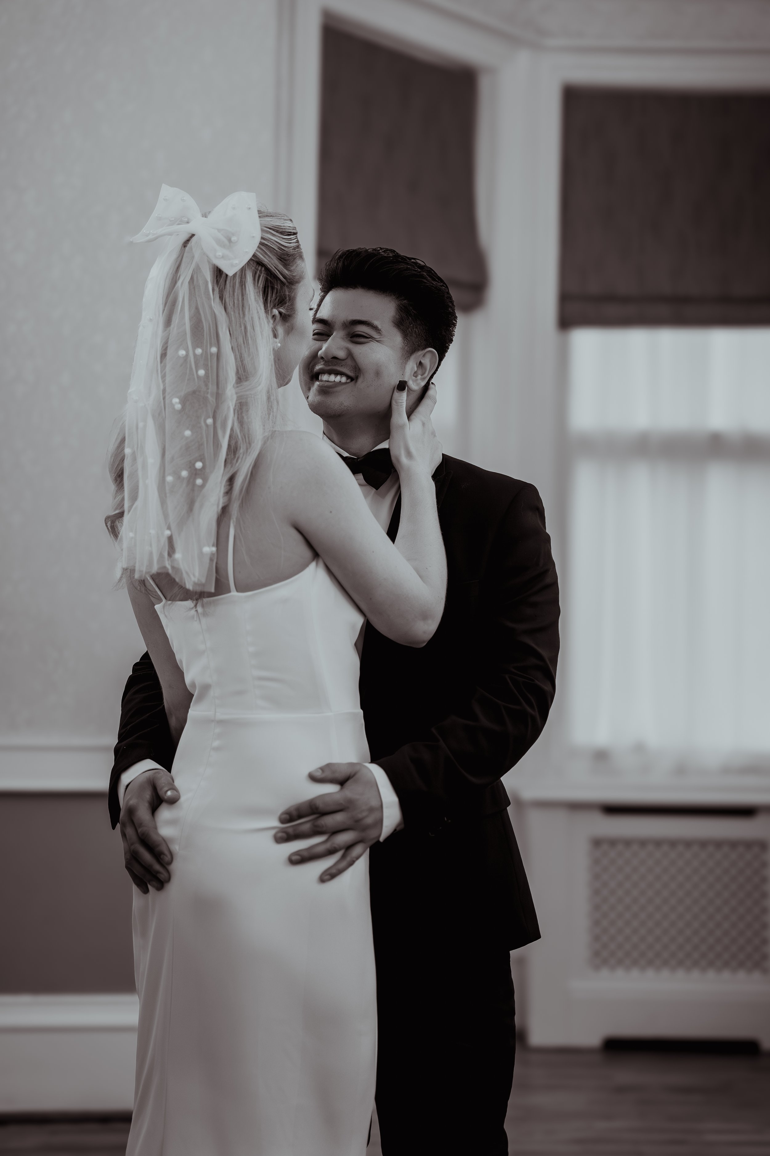 A bride and groom dancing intimately at their wedding, smiling and gazing at each other, indoors with large windows and curtains in the background.