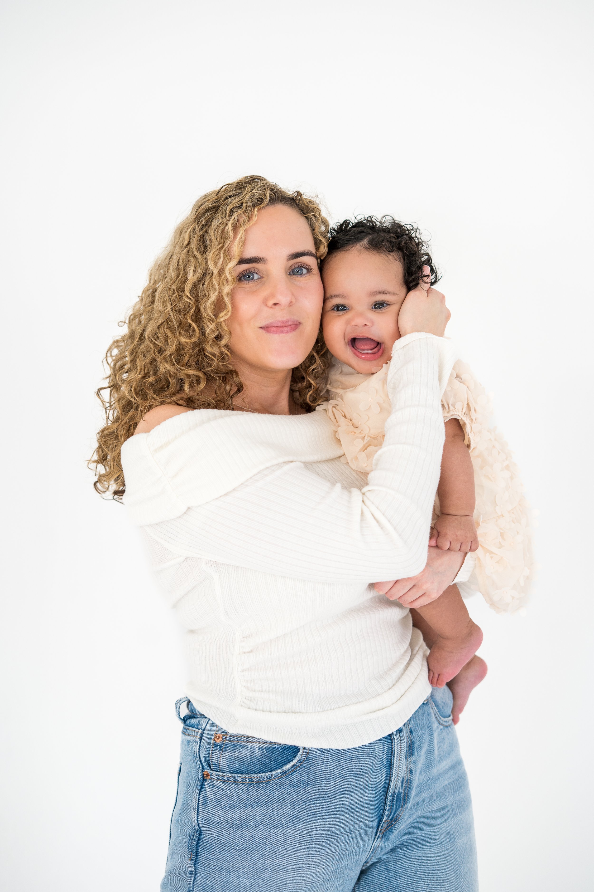 A woman with curly hair holding a young girl with dark curly hair and wearing a light-colored dress, both smiling and looking at the camera against a plain white background.