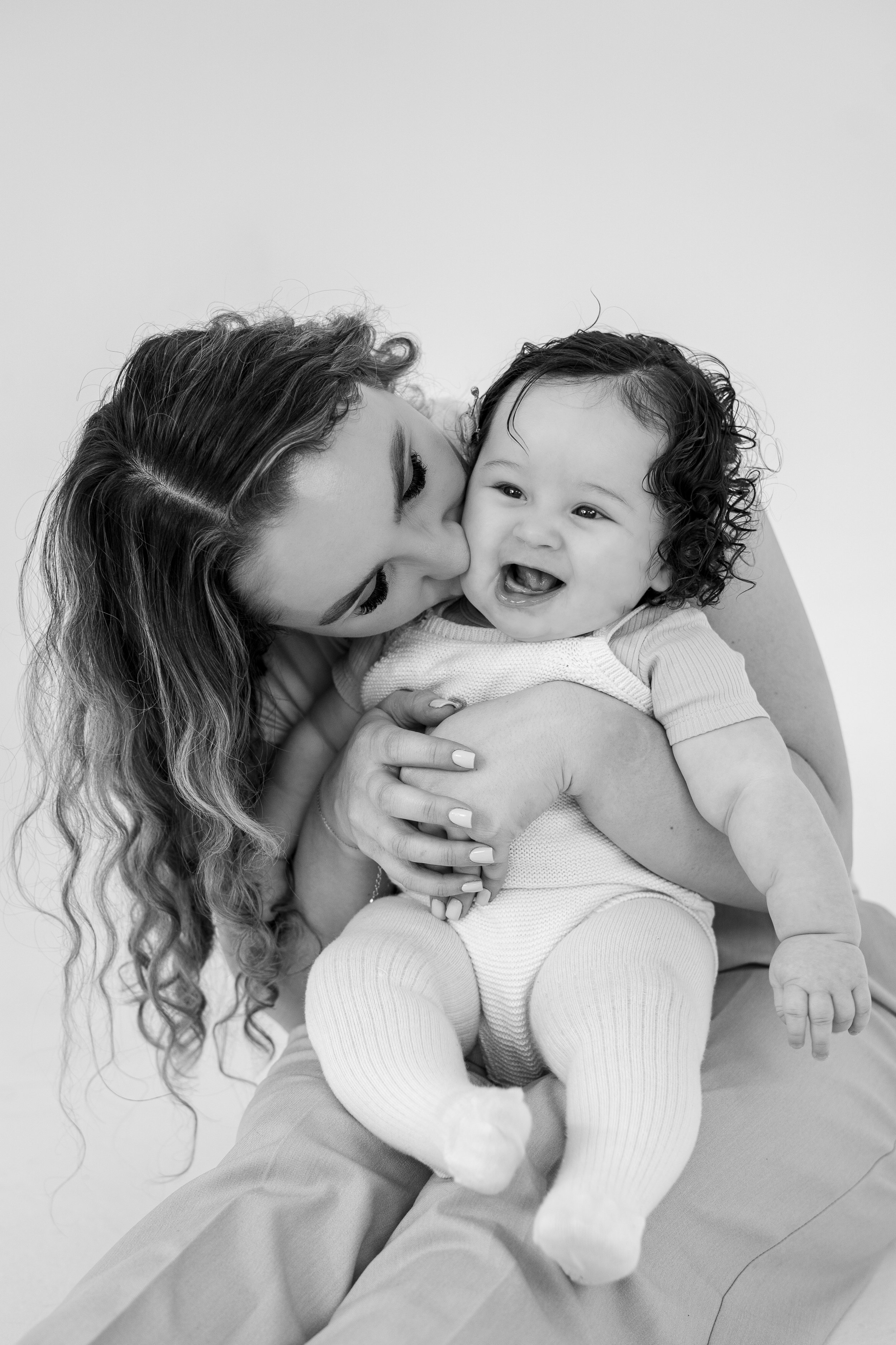 A woman is holding a smiling baby while giving the baby a kiss on the cheek