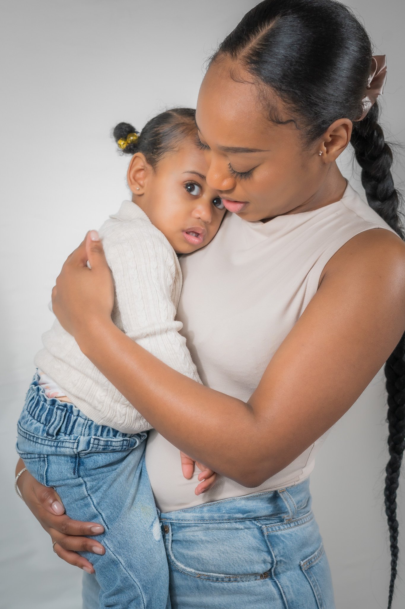 A woman with braided hair holding a young girl with pigtails close, both with neutral expressions, against a plain background.