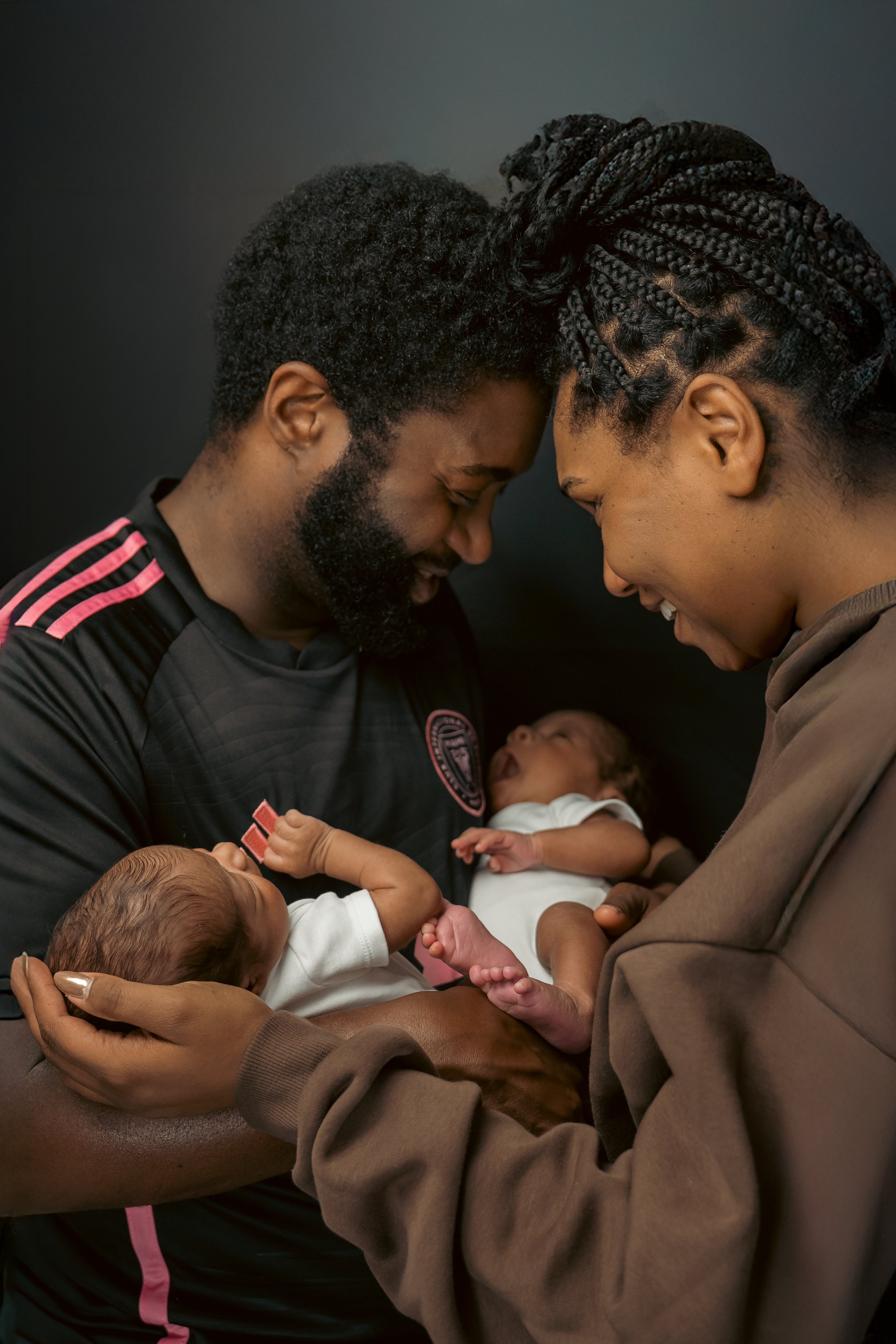 A man and woman, both smiling and touching foreheads, hold two newborn babies in their arms, looking at each other lovingly.