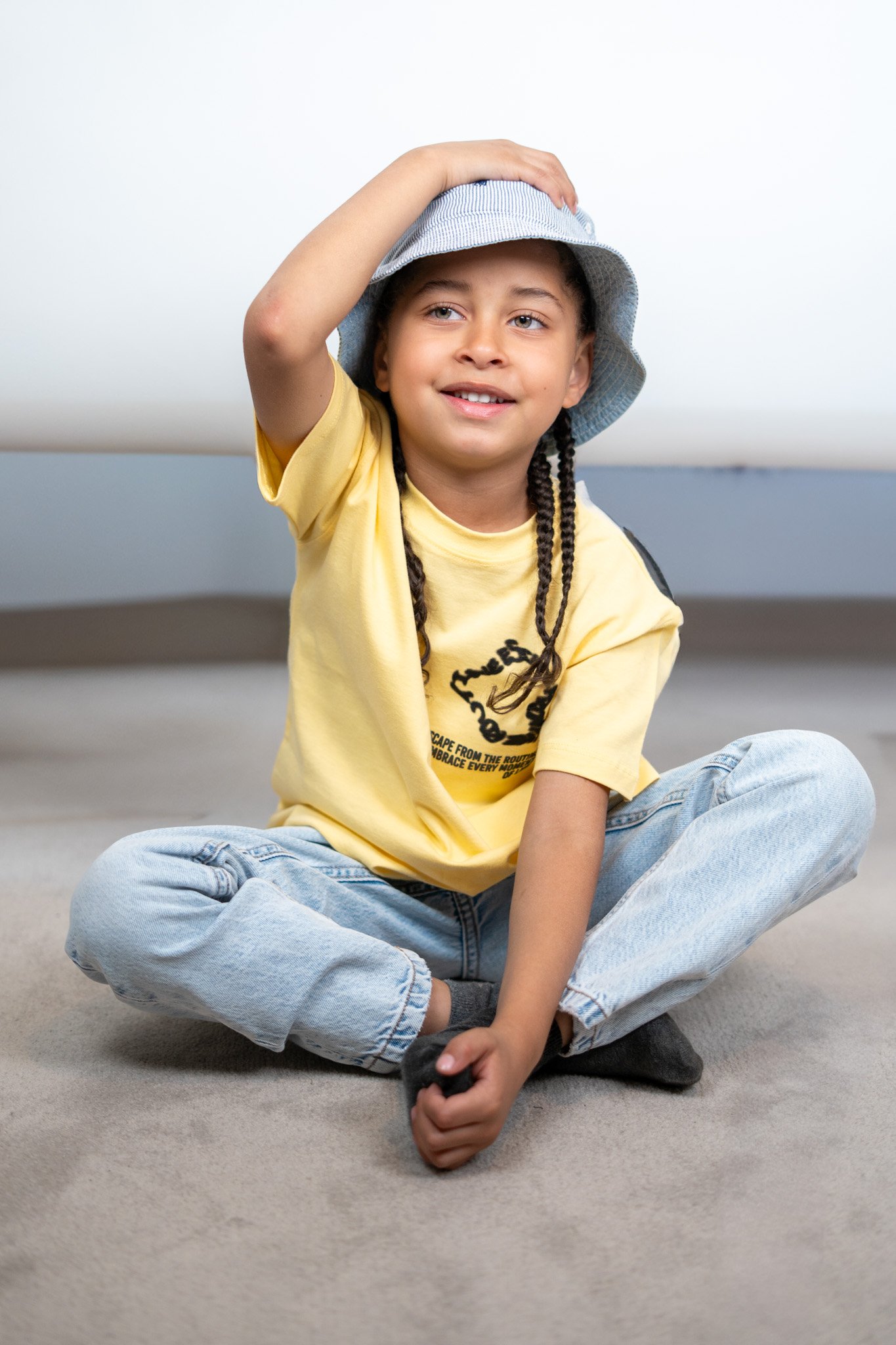 A young girl with braided hair, wearing a yellow T-shirt, light blue jeans, gray socks, and a blue striped bucket hat, sitting cross-legged on a carpeted floor, smiling, with her right hand on her head.