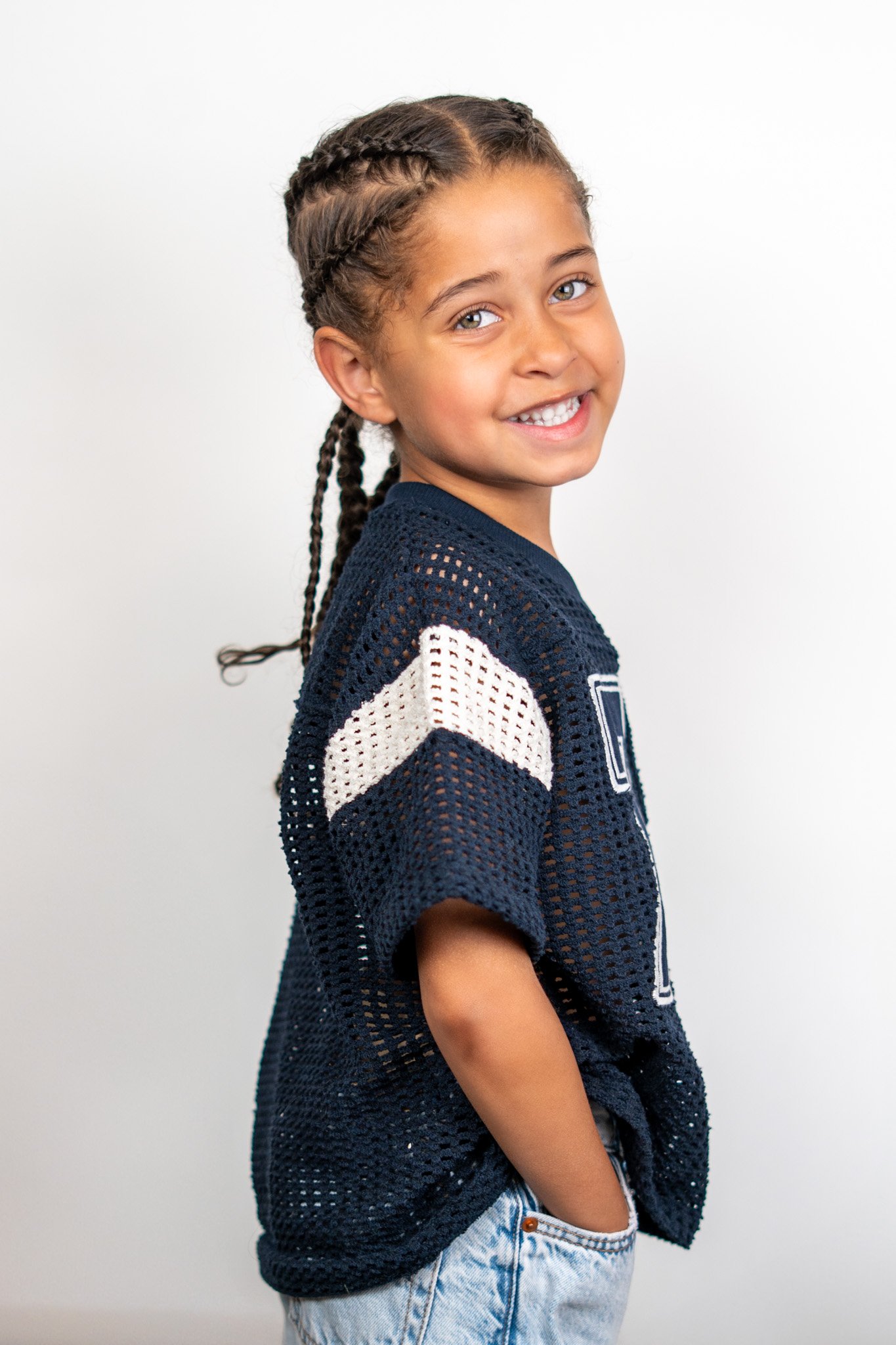 A young girl with braided hair smiling and wearing a navy blue open-knit sweater with white accents and light blue jeans, standing against a plain white background.