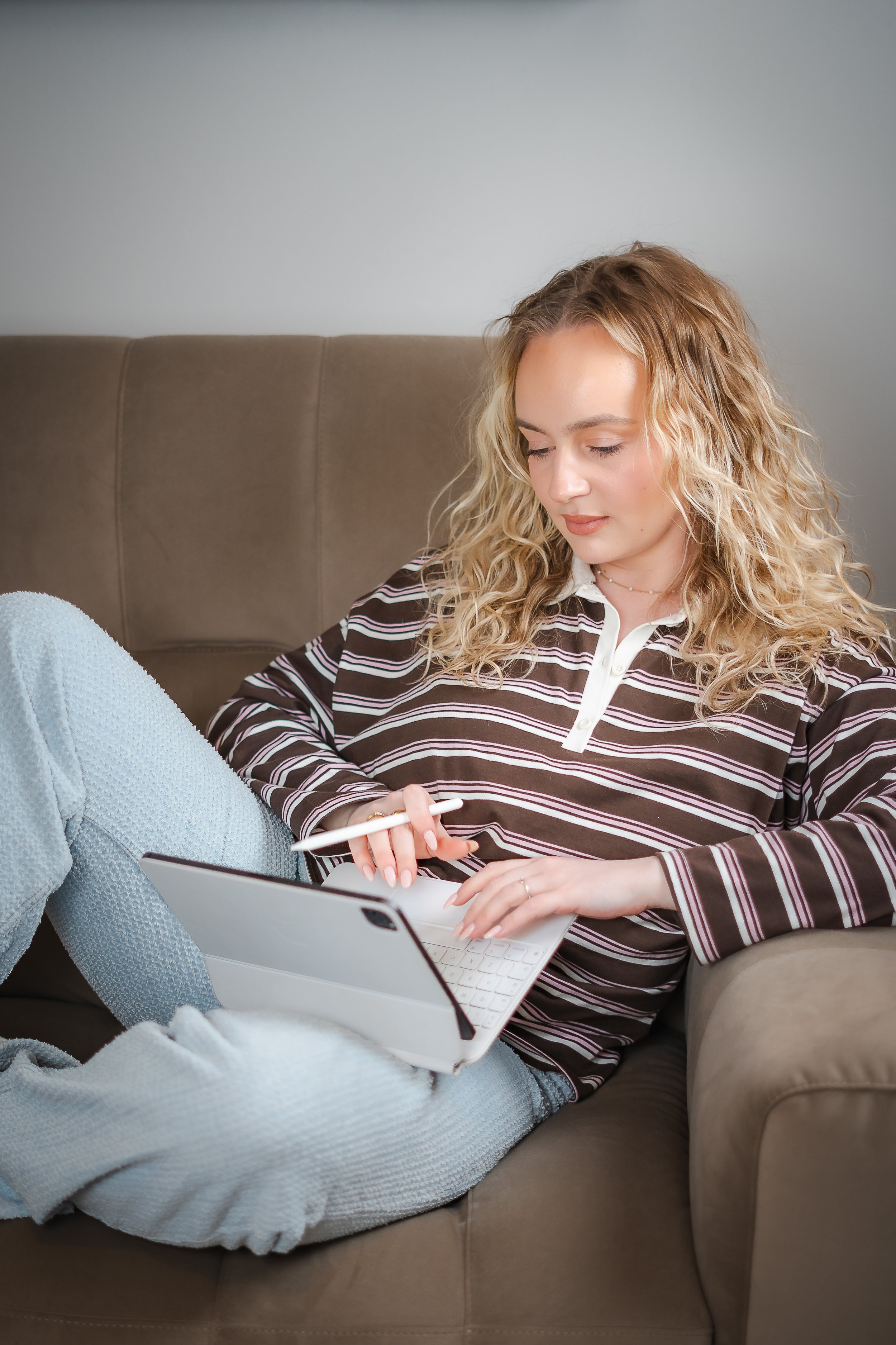 Young woman with curly blonde hair sitting on a brown sofa, using a tablet with a stylus, wearing a striped long-sleeve shirt and light blue textured pants.