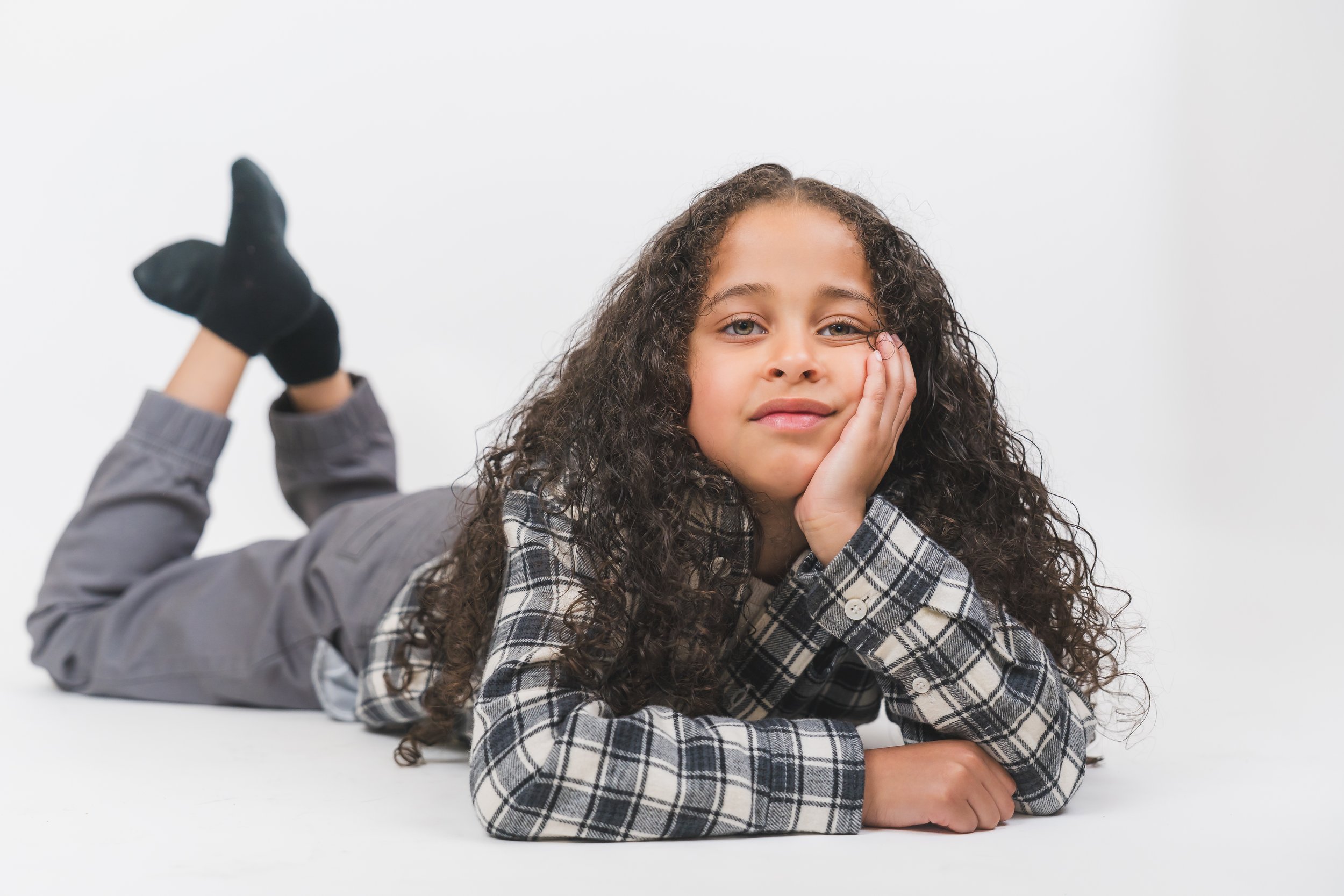 Young girl with curly hair lying on her stomach, resting her head on her hand, wearing a plaid shirt and gray pants, against a plain white background.