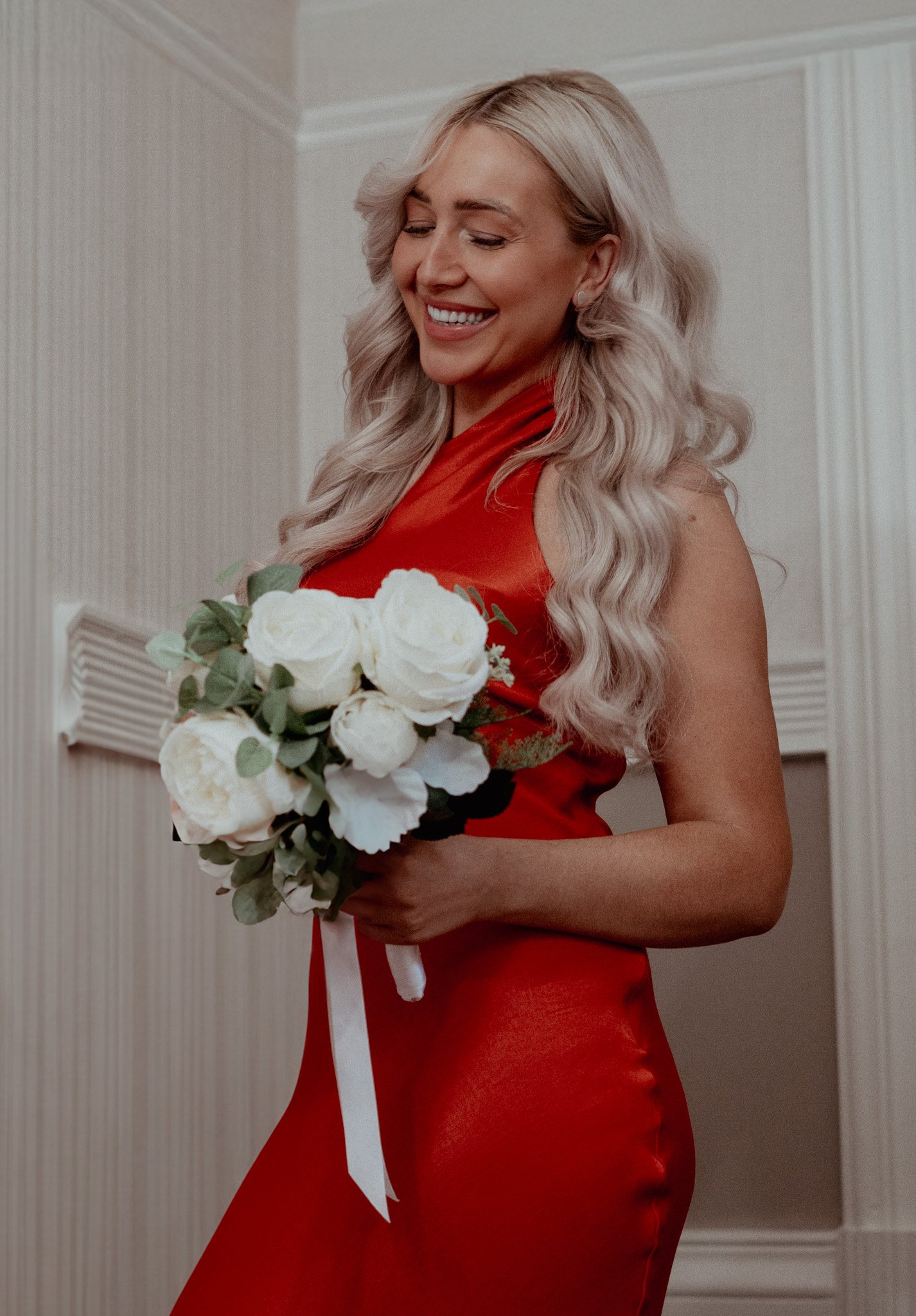 A woman with long blonde wavy hair wearing a red dress, smiling and holding a bouquet of white flowers with greenery, standing indoors with cream-colored walls and molding.