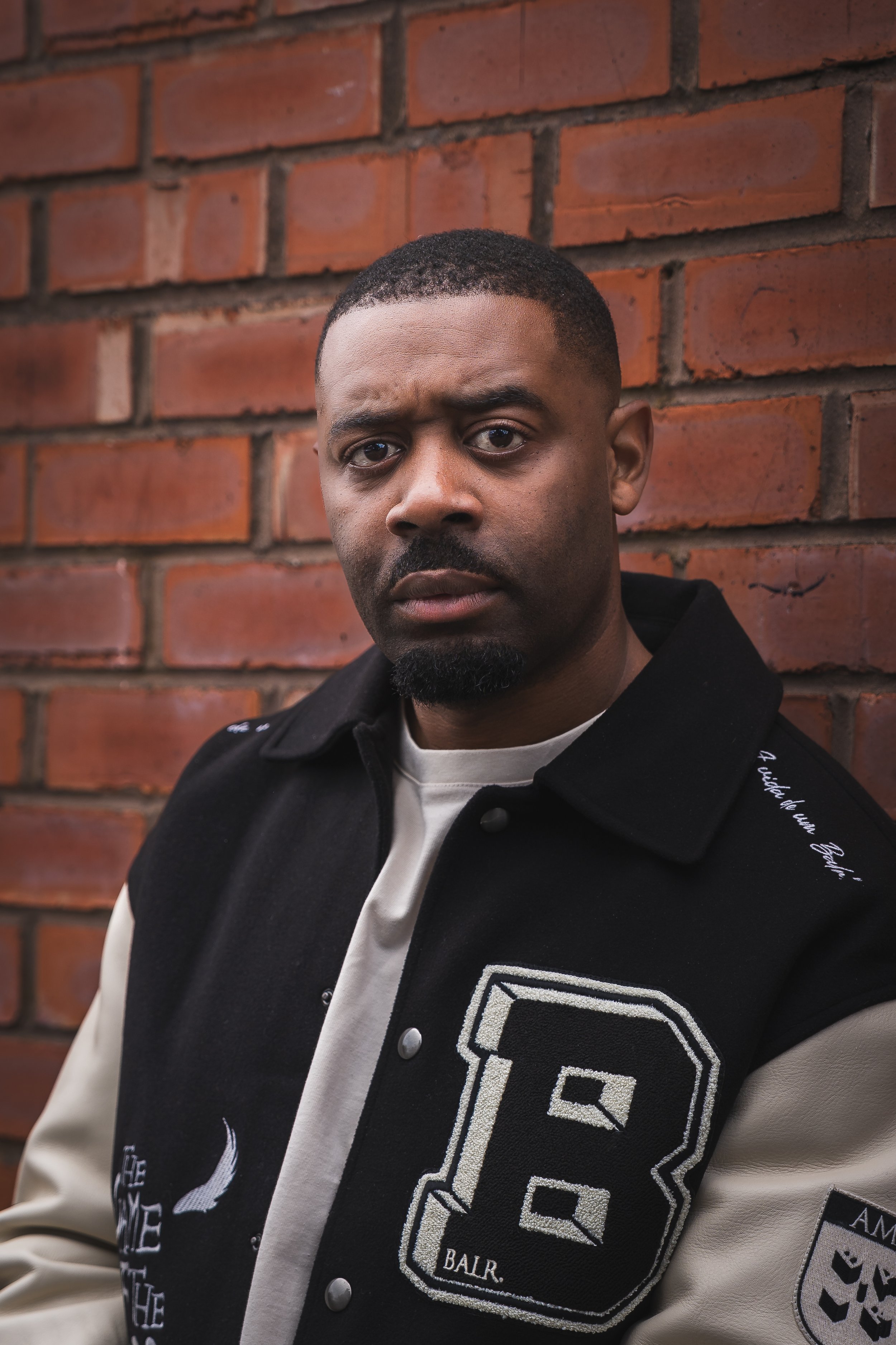 Portrait of a young man standing against a red brick wall, wearing a black and beige varsity jacket with patches.