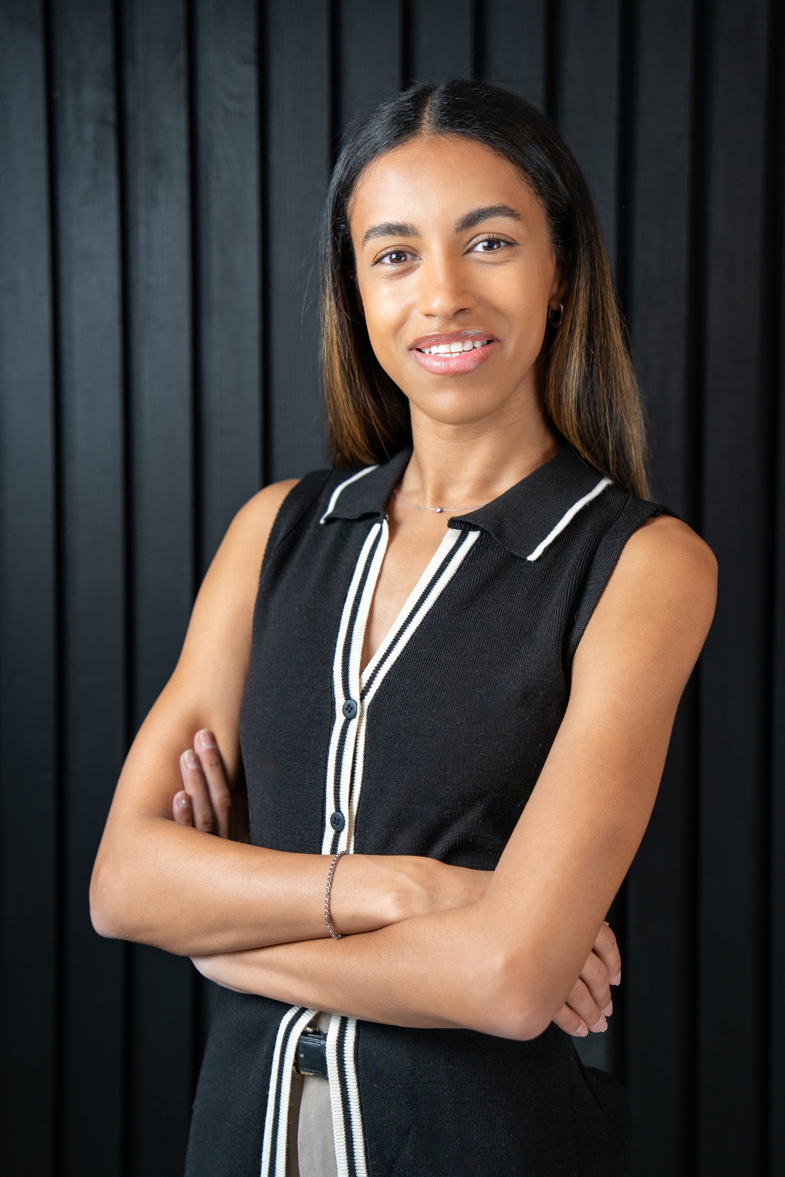 A young woman with medium skin tone and straight brown hair, wearing a sleeveless black dress with white trim, standing against a dark wooden background with her arms crossed, smiling slightly.