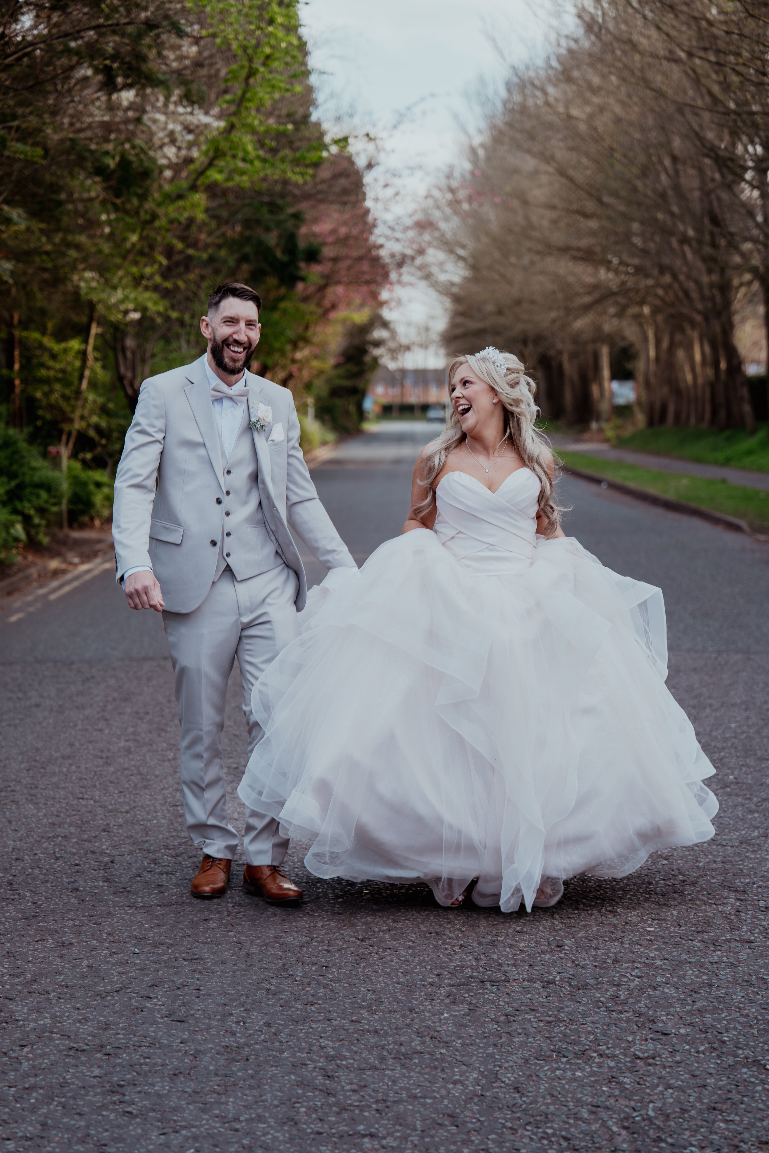 A bride and groom walking hand in hand on a street lined with trees, smiling and laughing during their wedding day.