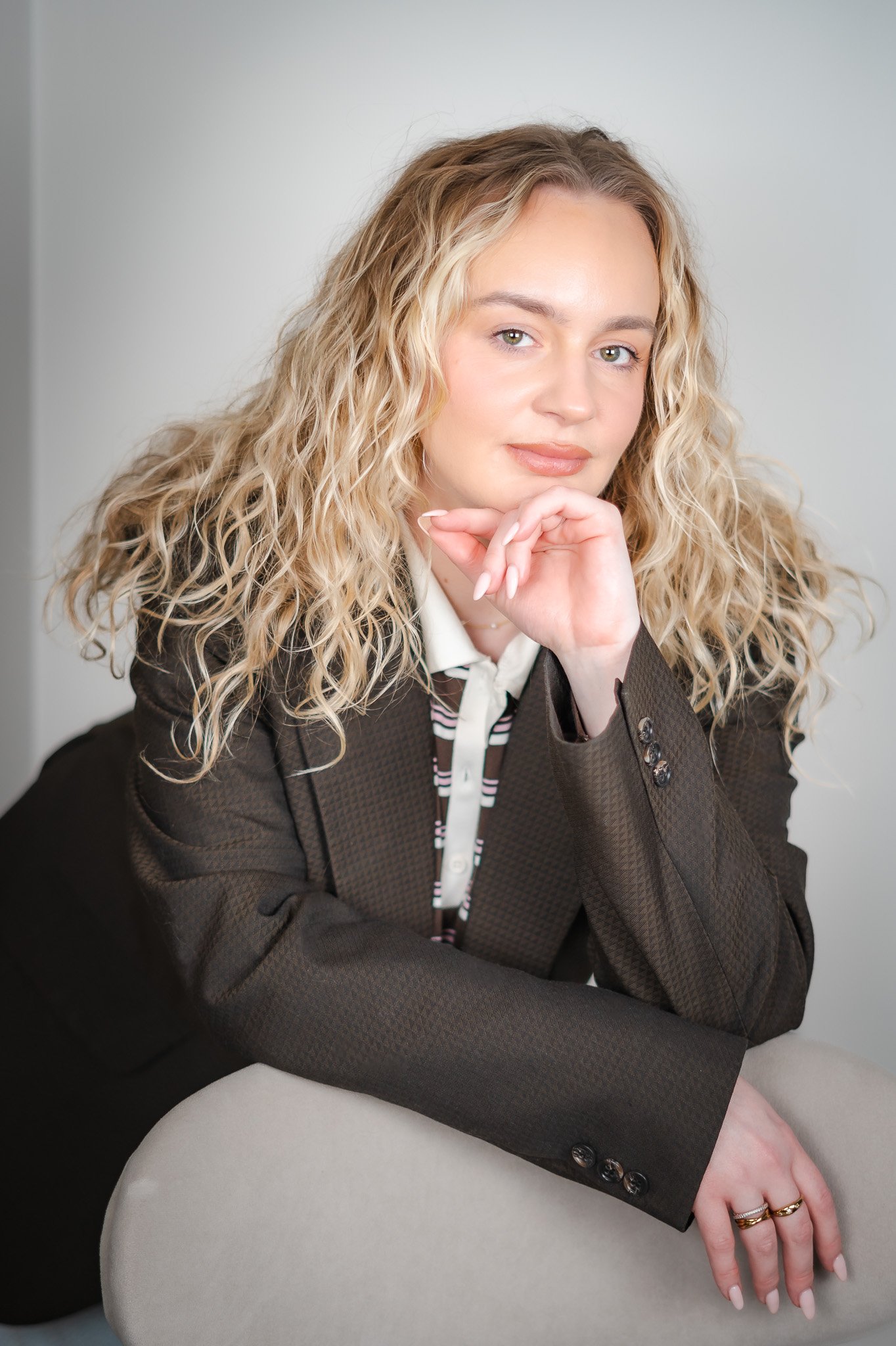 A woman with blonde, curly hair rests her chin on her hand, wearing a dark blazer and a striped shirt, sitting on a light-colored chair.