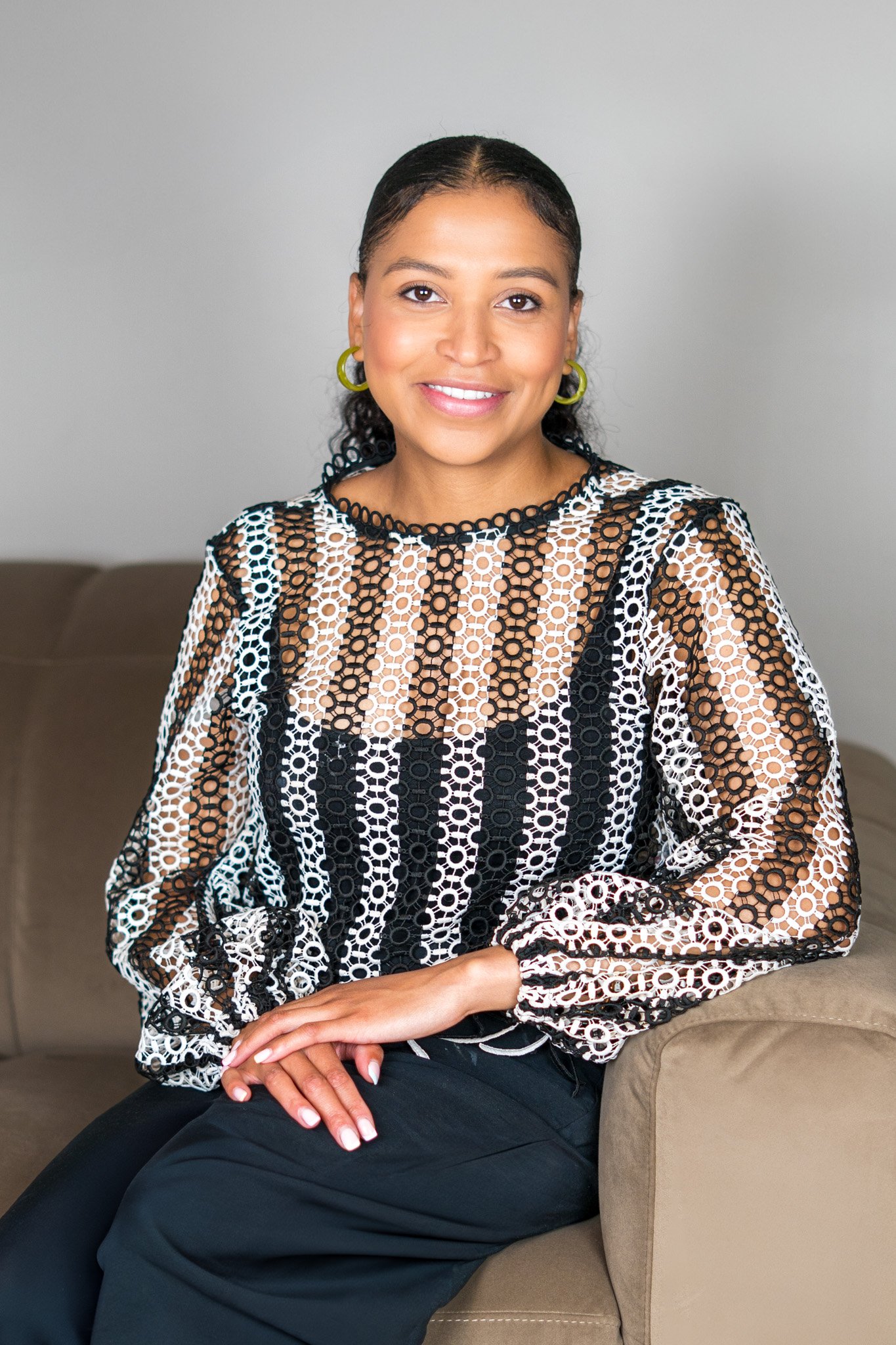 A woman with dark hair pulled back, wearing a black and white patterned long-sleeve blouse and black pants, sitting on a beige couch with a neutral background, smiling at the camera.