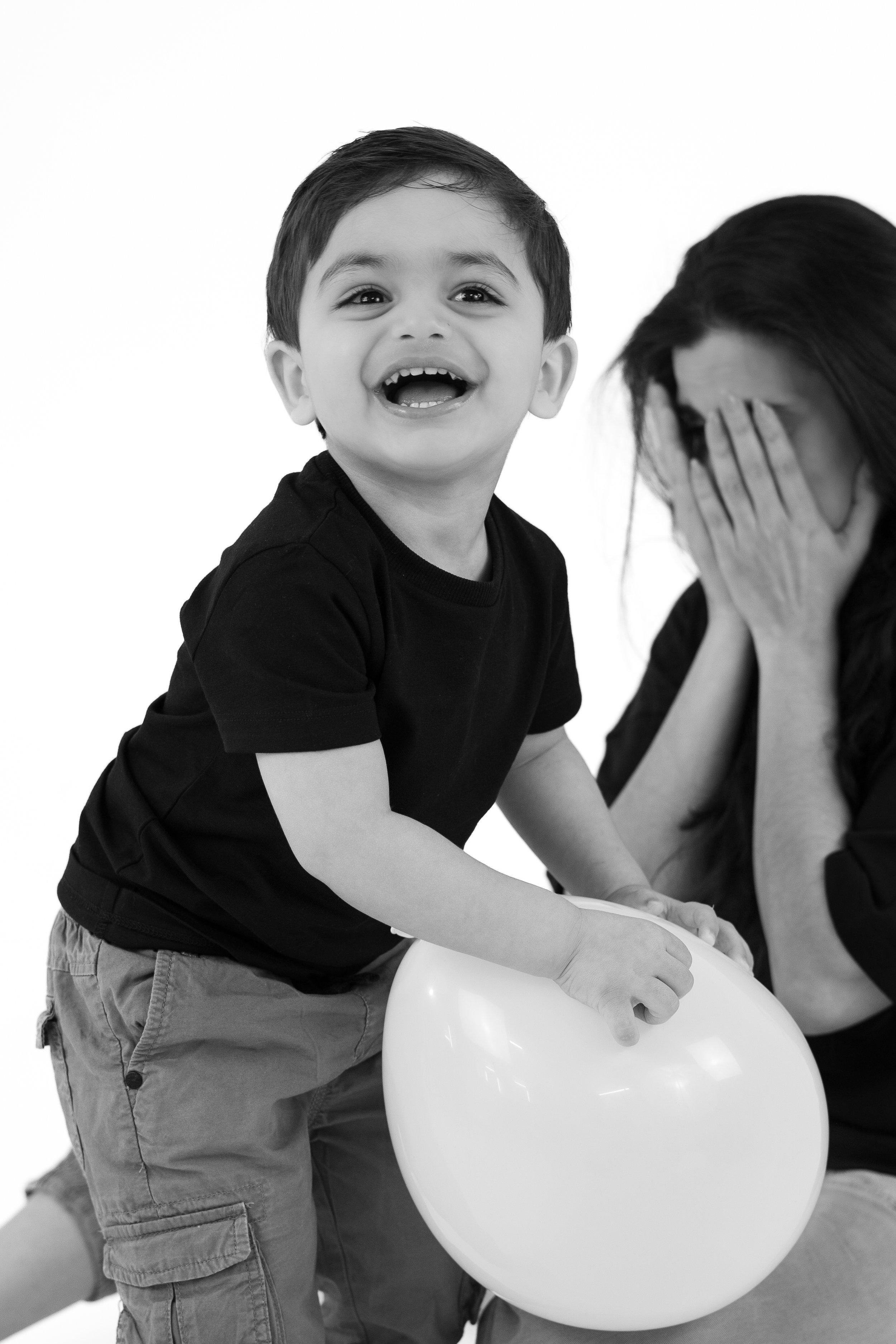 A young boy laughing and holding a balloon while a woman sits beside him covering her face with her hands.