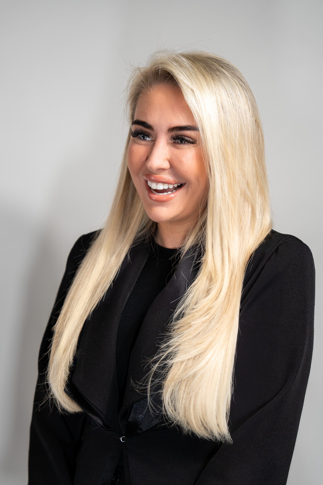 A woman with long blonde hair, wearing a black blazer, smiling and looking to her left, against a light gray background.