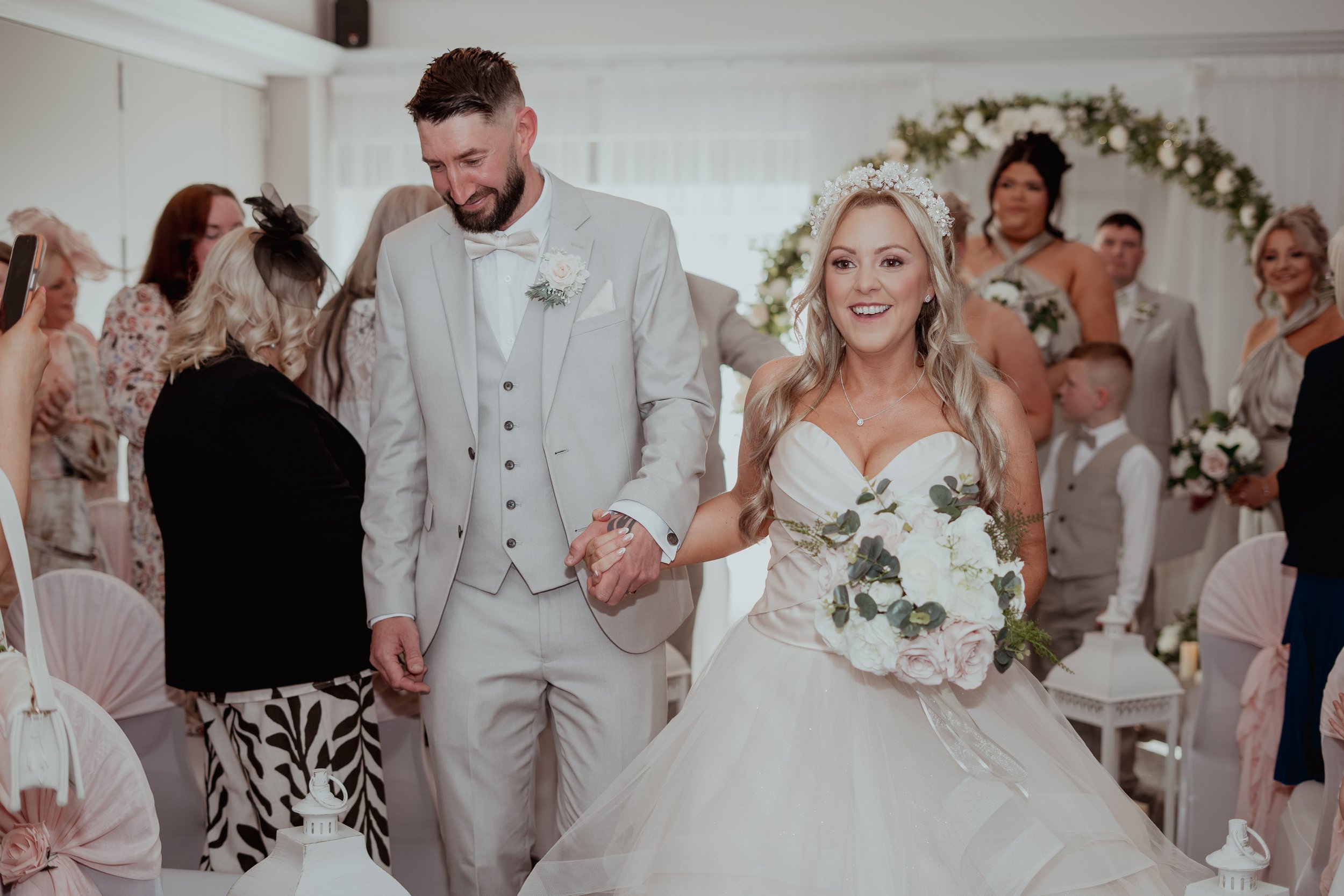 A bride and groom holding hands and walking down the aisle at their wedding ceremony, surrounded by guests. The bride is wearing a white wedding dress and holding a floral bouquet, smiling, and wearing a flower crown. The groom is wearing a light-col