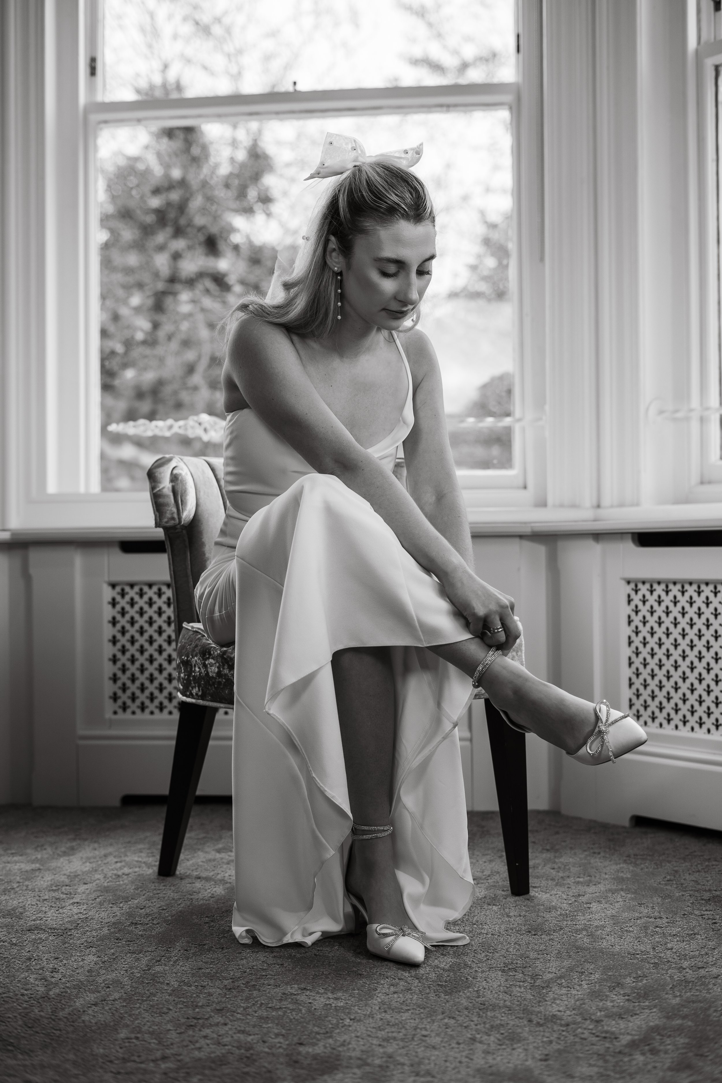 A woman in a wedding dress sitting on a chair, adjusting her shoe, in front of a window, in black and white.