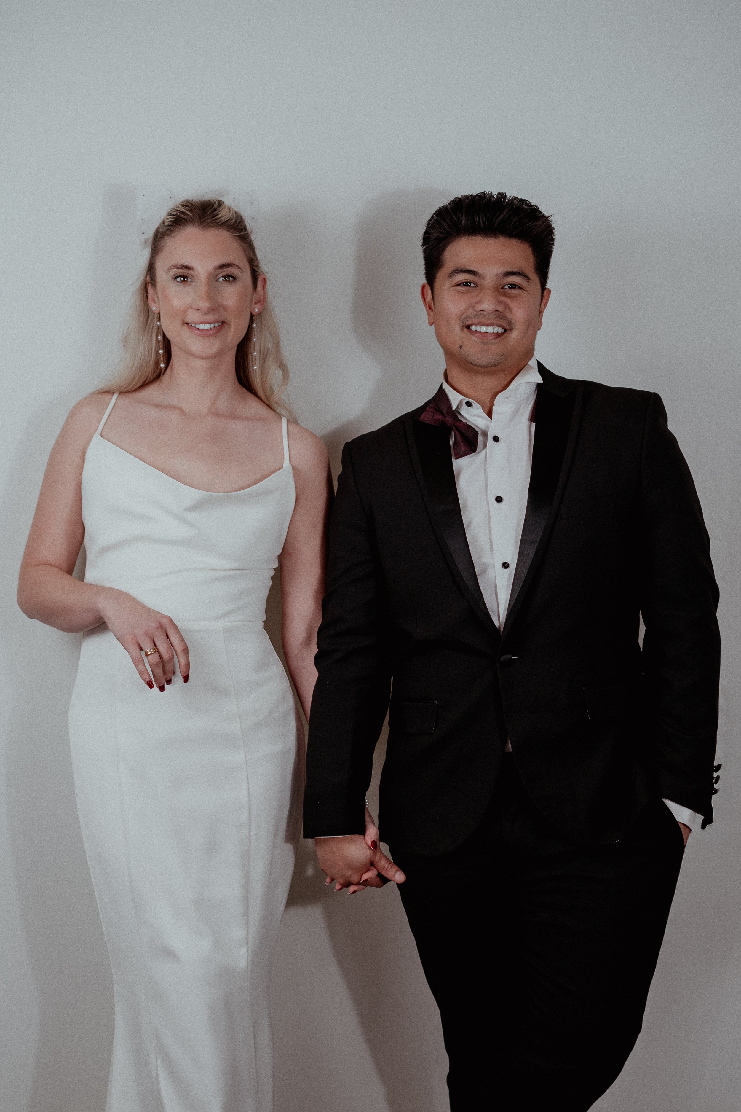 A couple in formal attire holding hands and smiling, standing against a plain white wall.