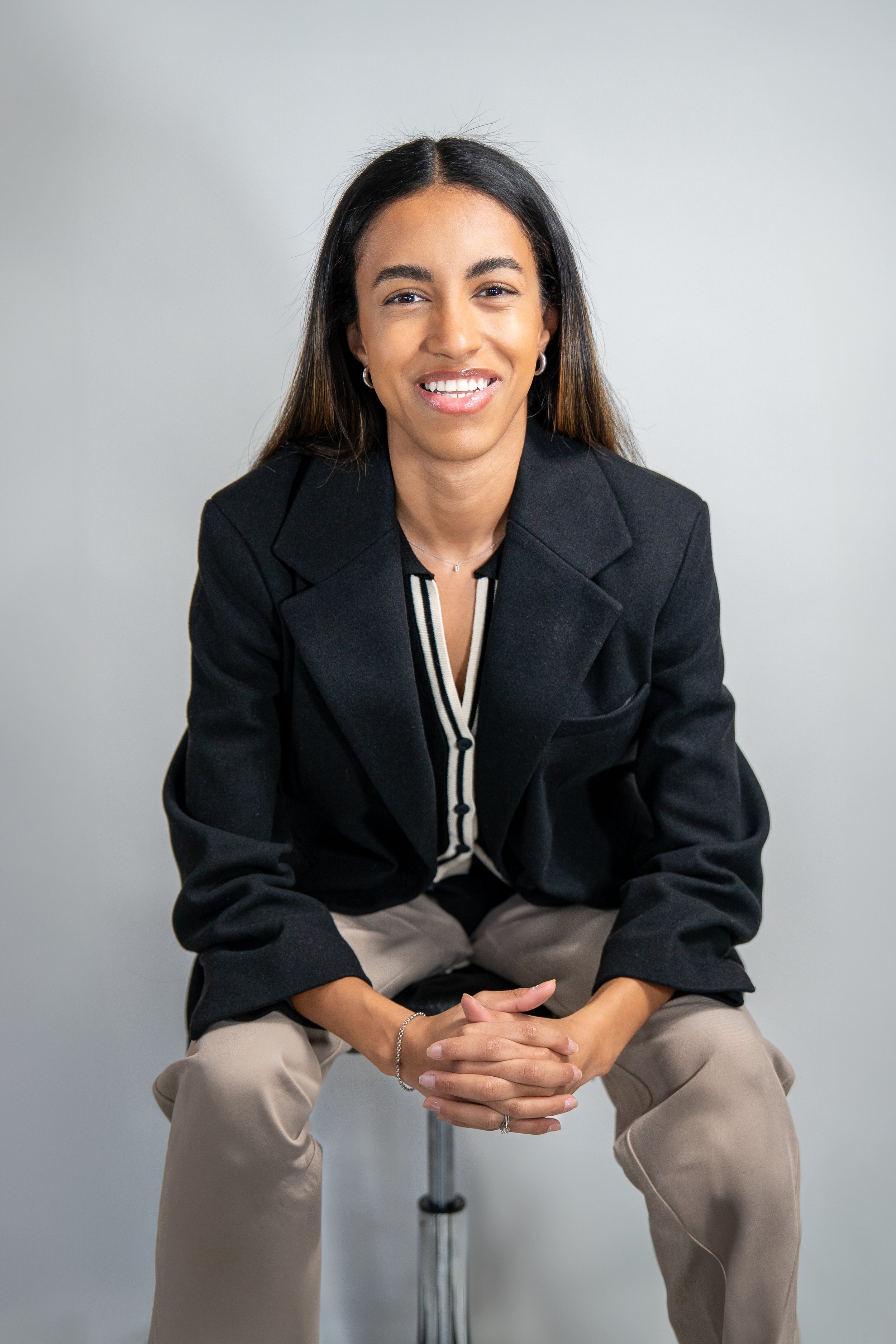 A woman with long dark hair wearing a black blazer and beige pants, sitting on a stool against a plain light background.