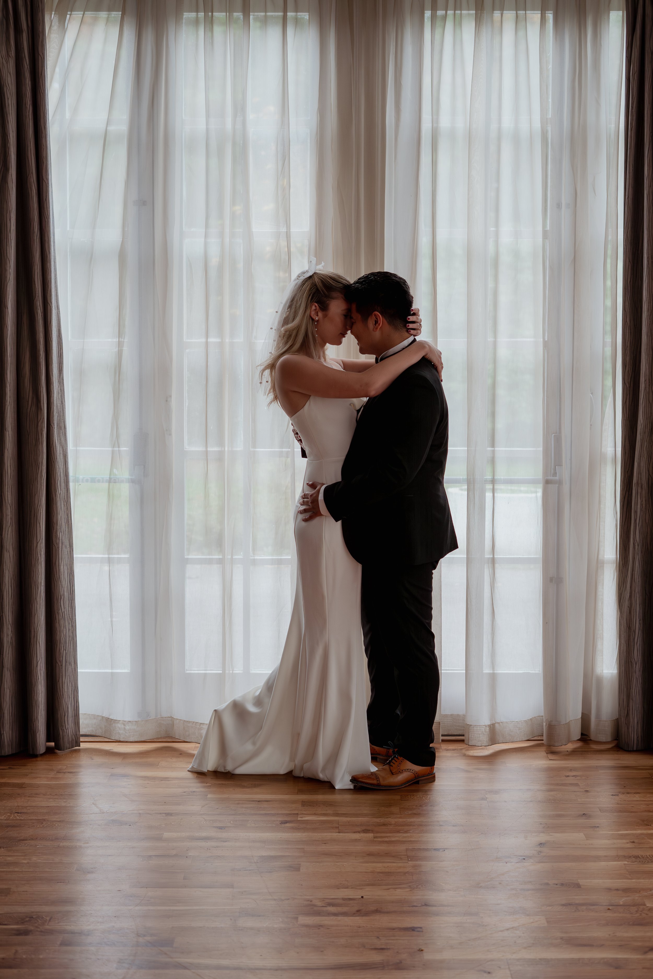 A bride and groom embracing in front of a large window with sheer curtains, backlit, on a wooden floor.