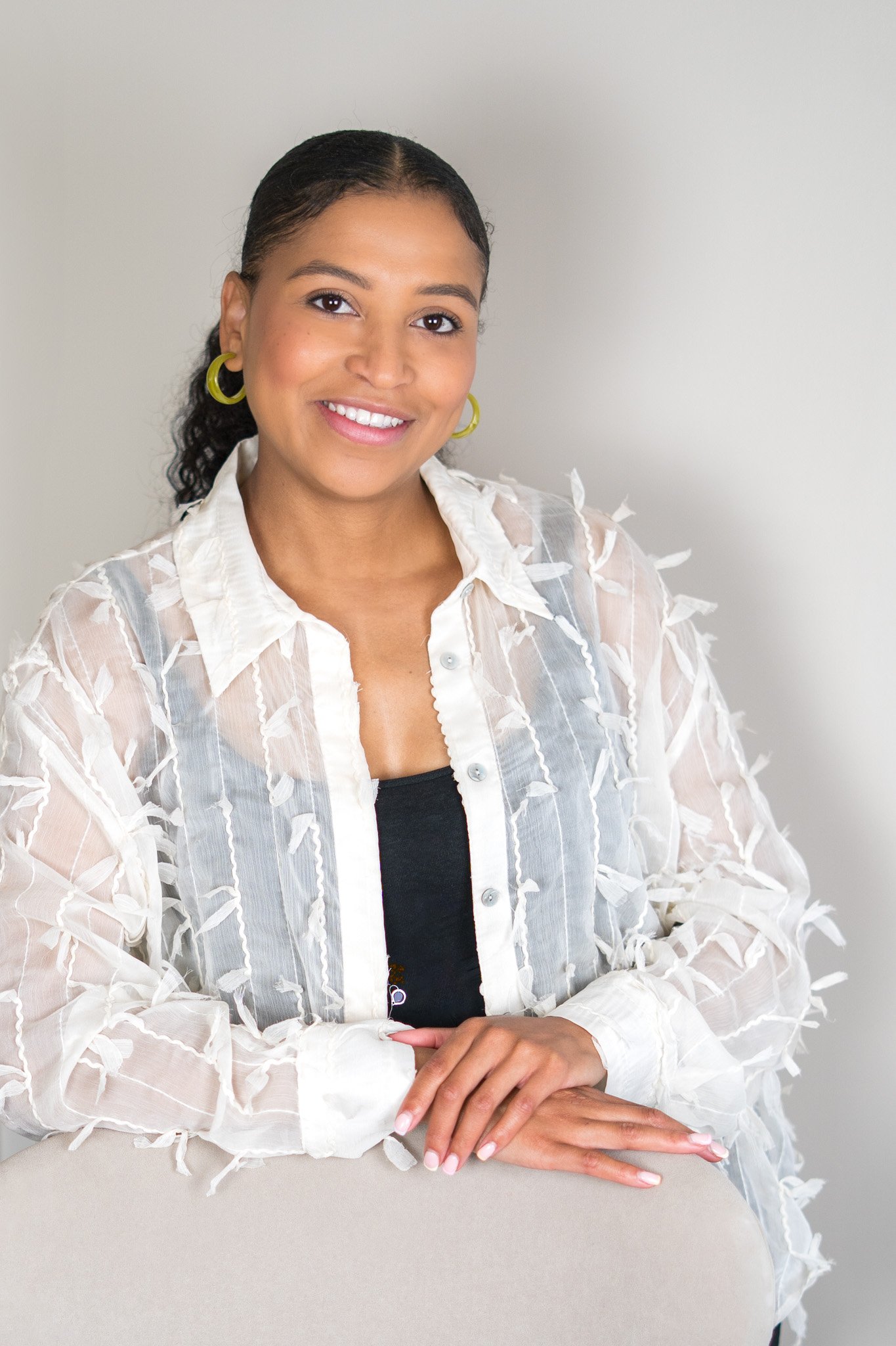 A woman with dark hair, wearing a sheer white blouse with feather-like details and hoop earrings, smiling at the camera.