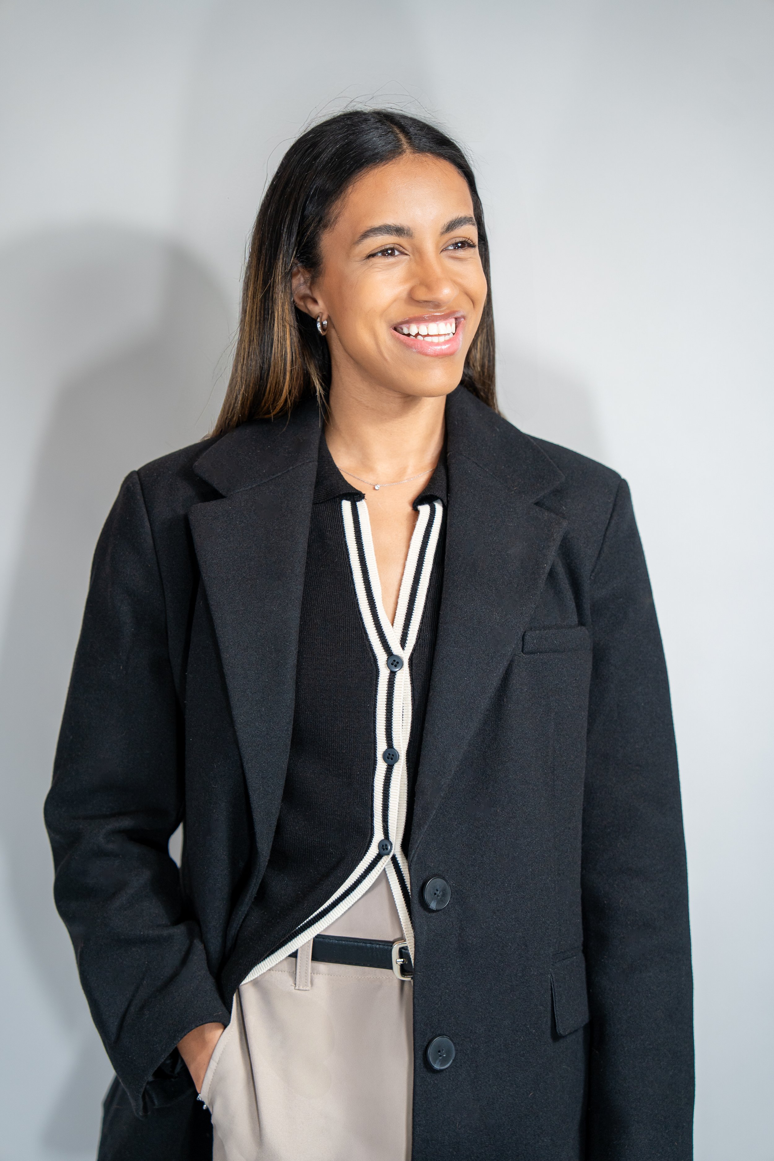 A smiling woman with long dark hair wearing a black blazer over a black and white striped blouse, beige pants, and a delicate necklace.
