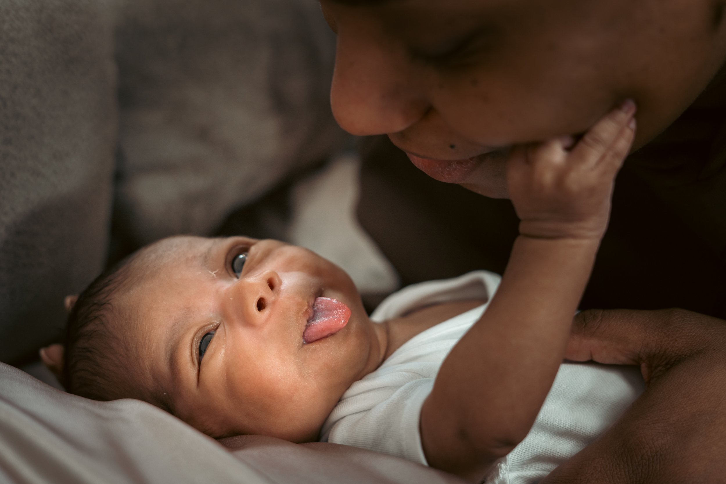 Close-up of a father and baby lying face-to-face, with the baby looking up at the father and sticking out its tongue, holding the father's chin with one hand.