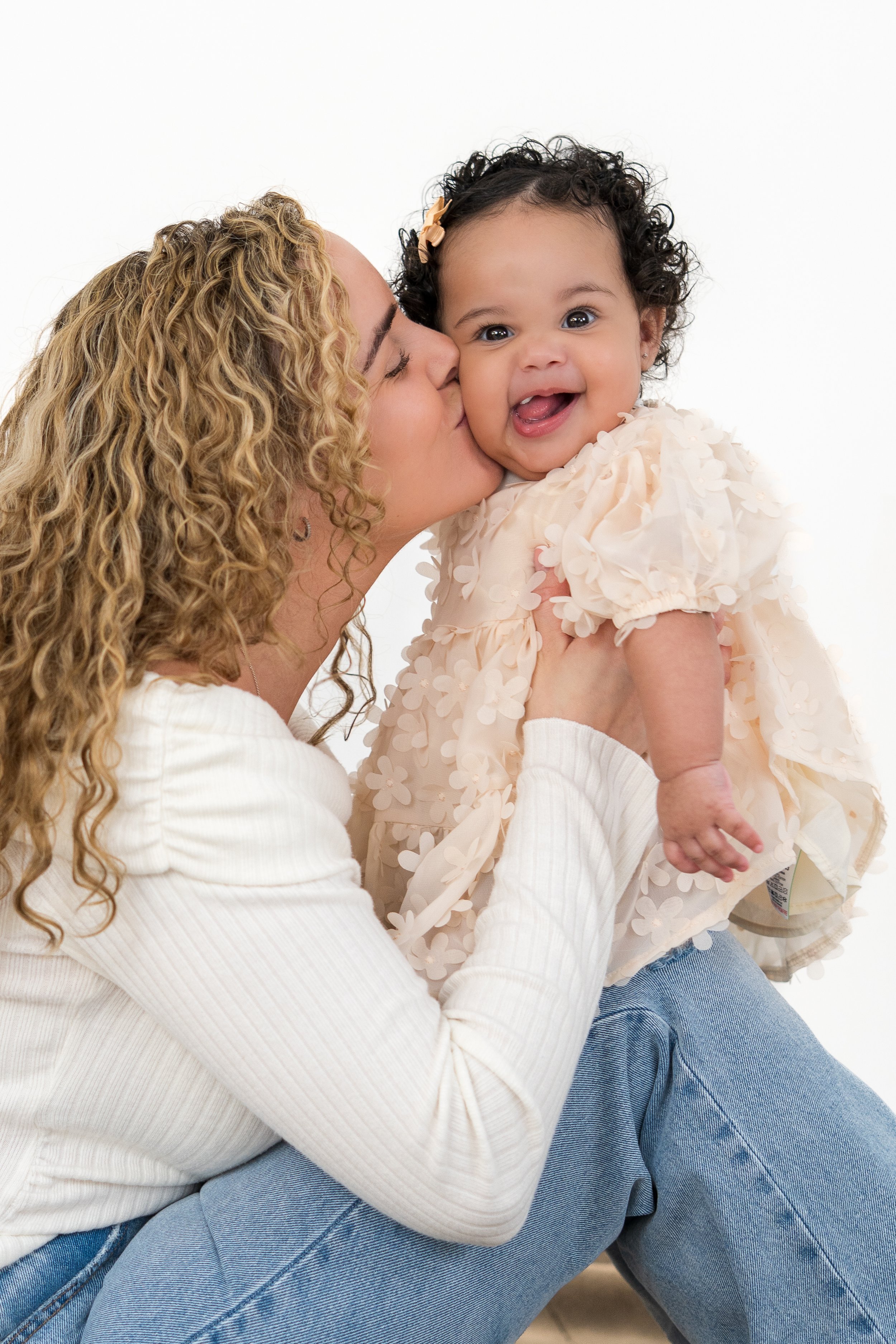 A woman with curly blonde hair kissing a smiling baby girl with dark curly hair, wearing a cream-colored dress with floral details, against a plain white background.