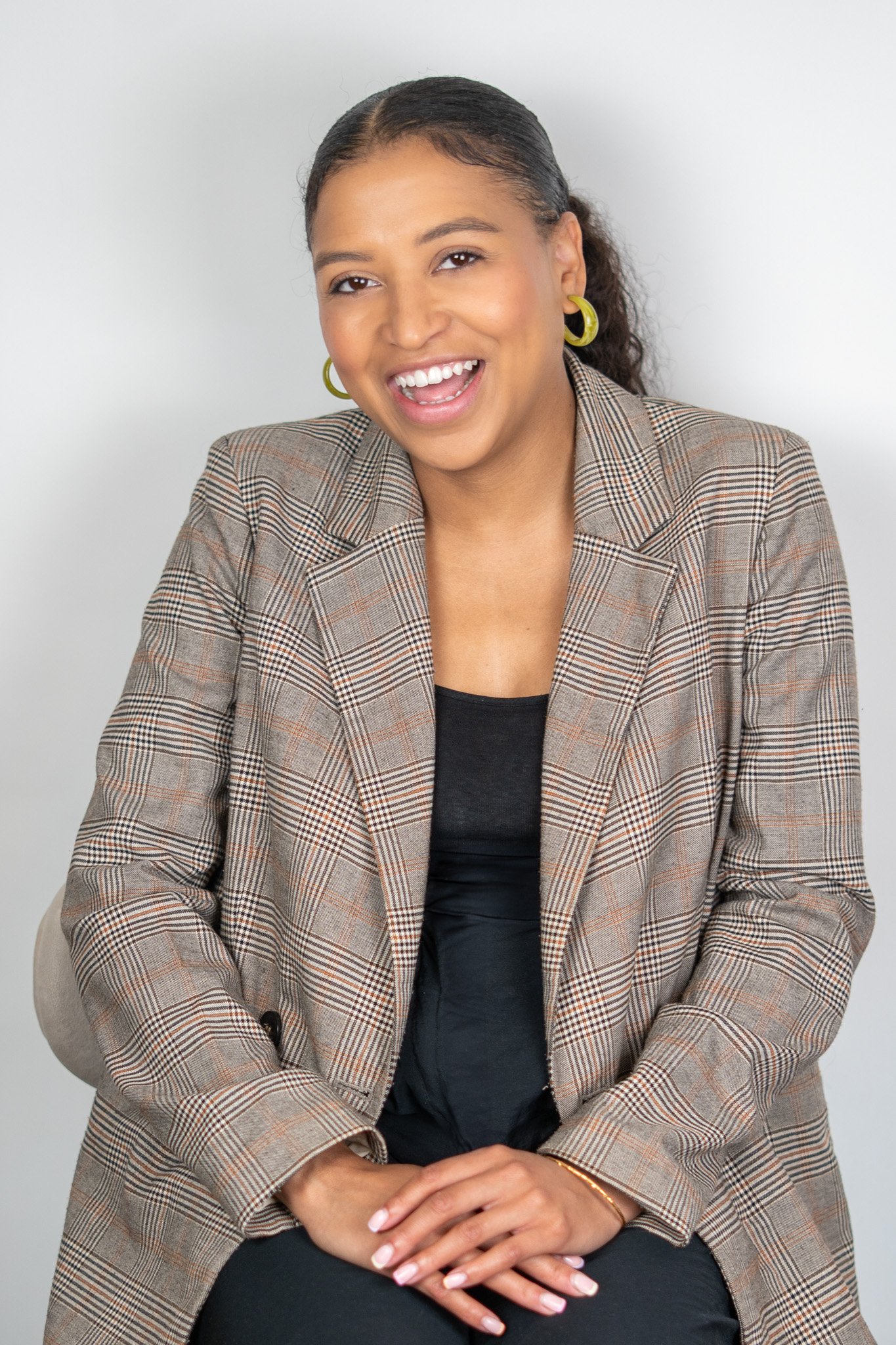 A smiling woman with dark hair tied back, wearing gold hoop earrings, a checkered blazer, and a black top, sitting against a plain white background.