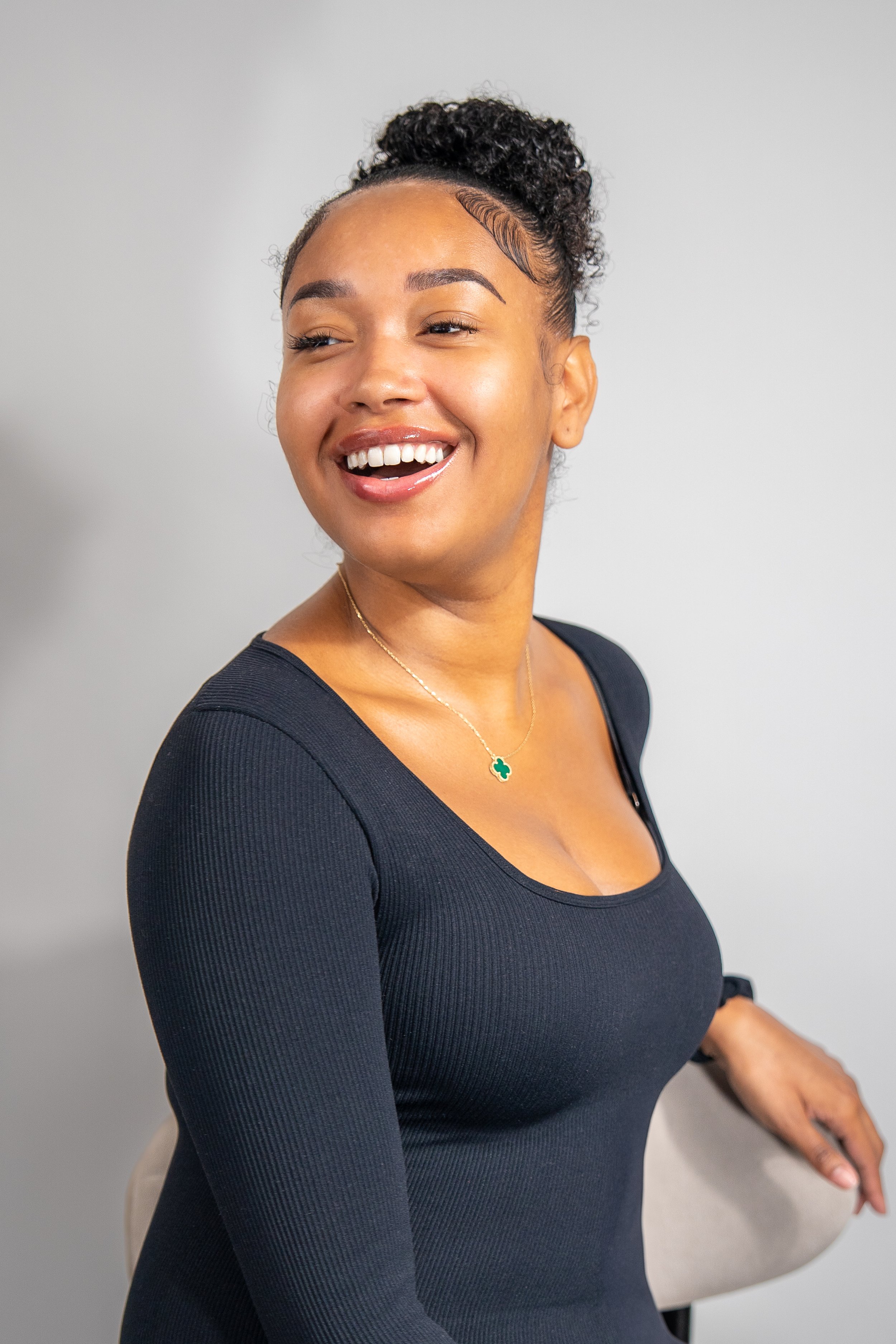 A smiling woman with curly hair tied up, wearing a black long-sleeve shirt and a necklace, looking to her left, seated against a neutral background.