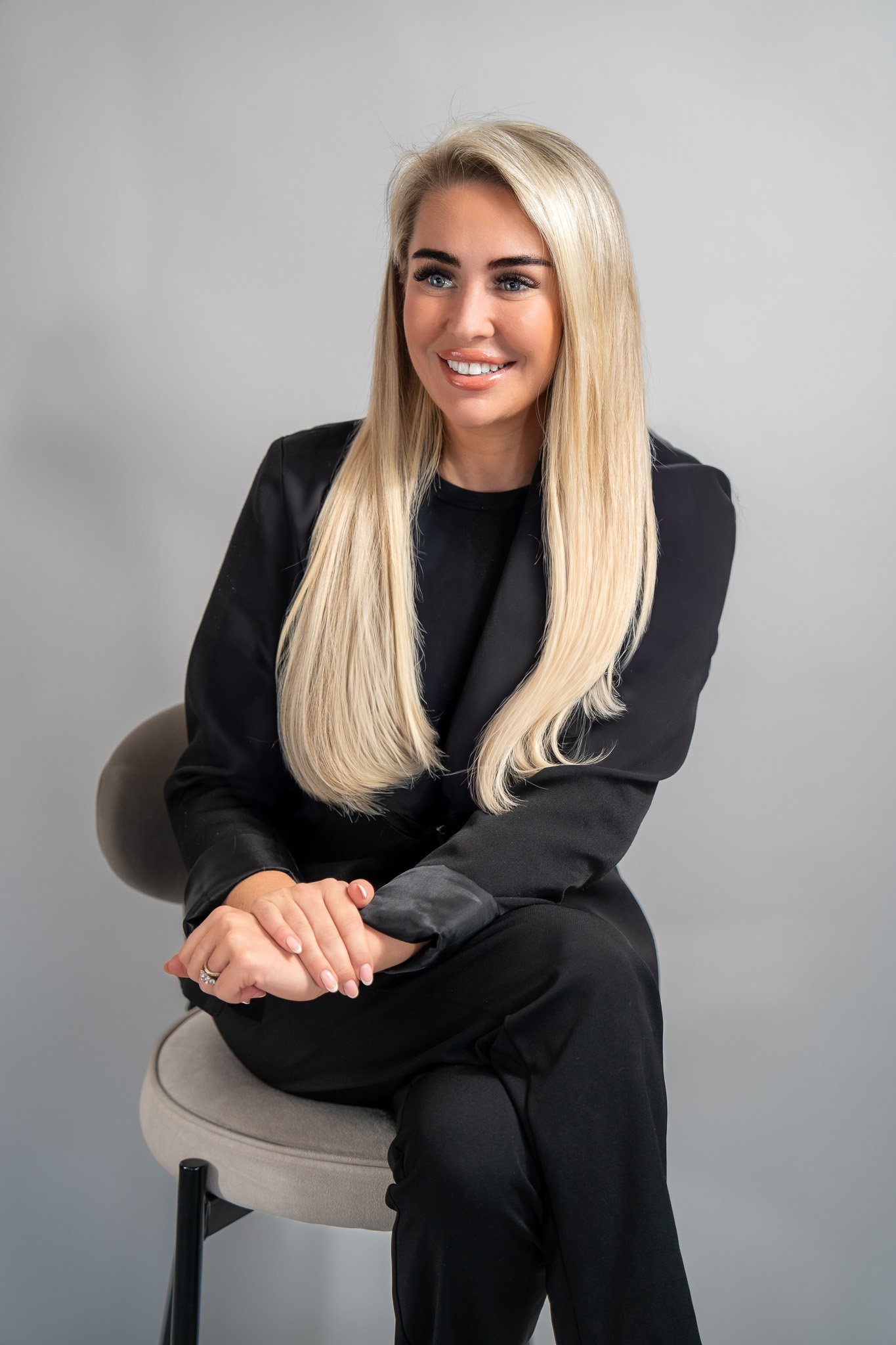 A woman with long blonde hair, wearing a black suit, sitting on a grey chair against a plain grey background, smiling and looking to the side.