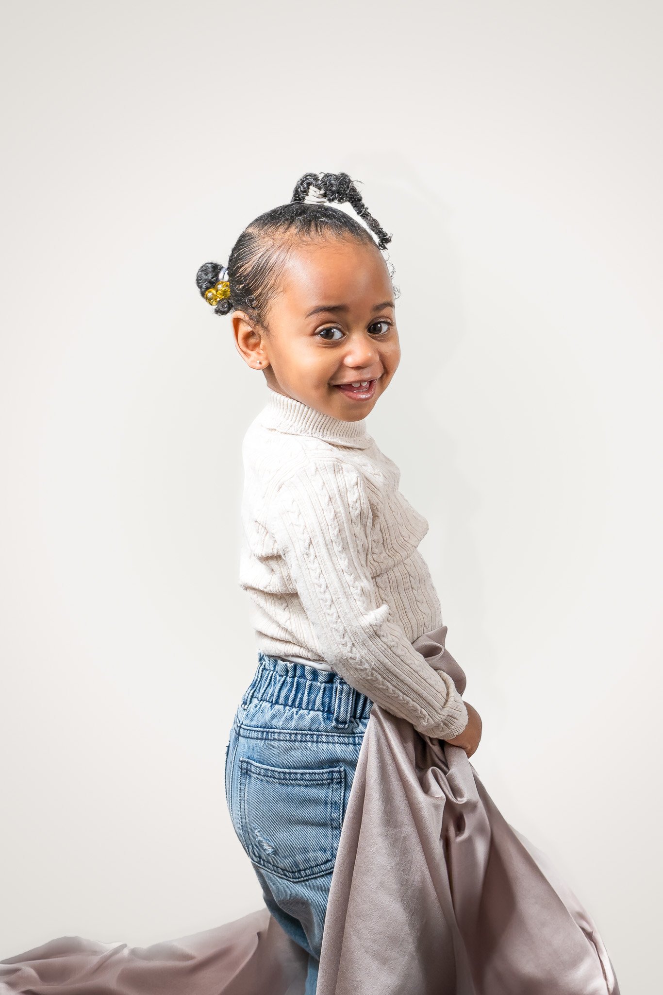 A young girl with pigtails wearing a white sweater and jeans, holding a piece of satin fabric, standing against a plain light-colored background and smiling at the camera.