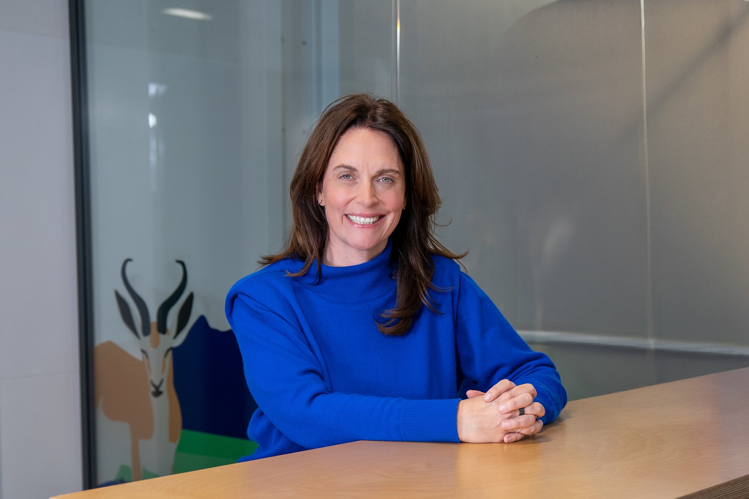 A woman with shoulder-length brown hair smiling while sitting at a wooden table indoors, wearing a bright blue sweater. Behind her, there is a glass wall with a mountain and antelope logo or design.
