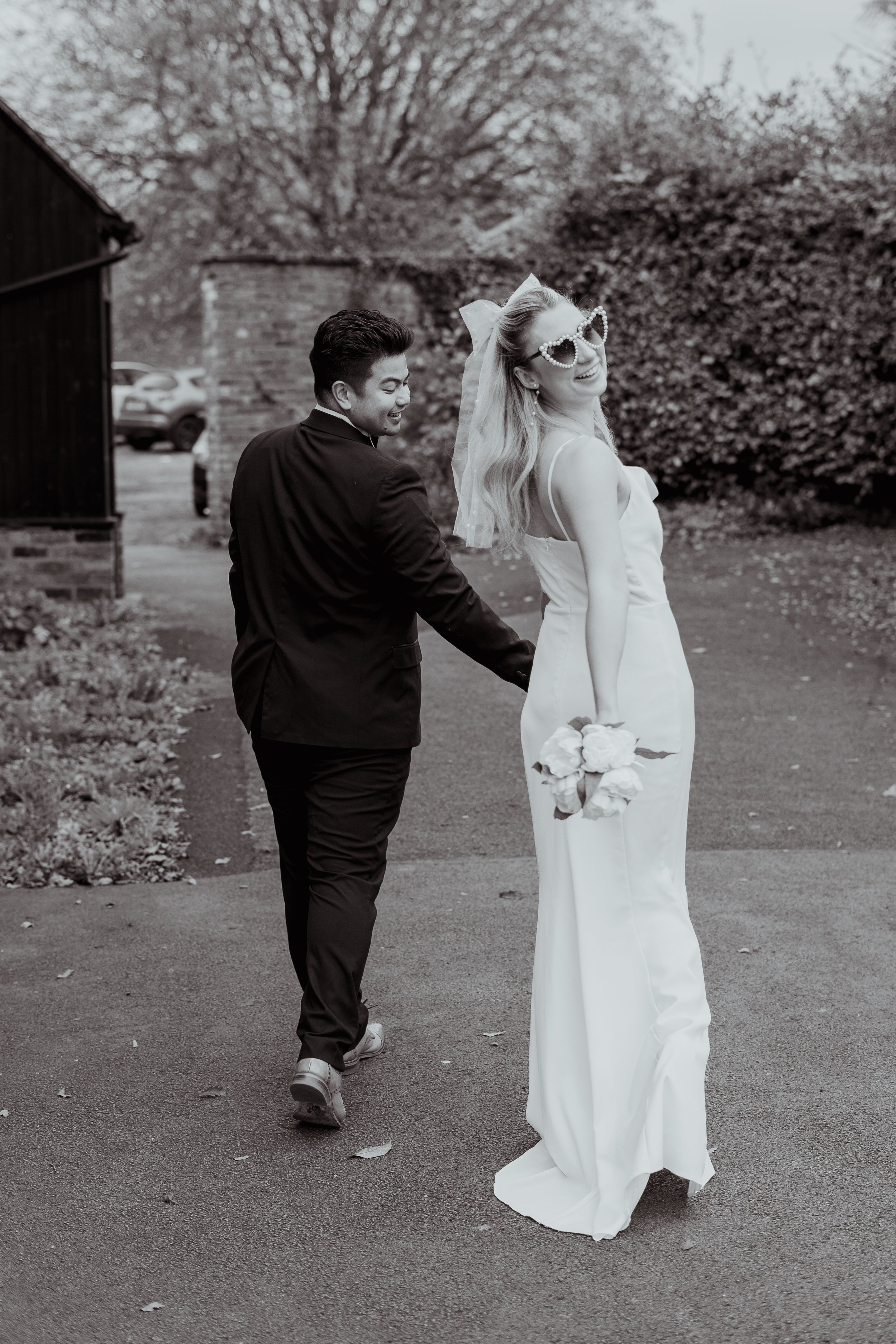 A bride and groom holding hands and walking outdoors, with the bride smiling and wearing heart-shaped sunglasses and holding a bouquet of flowers.