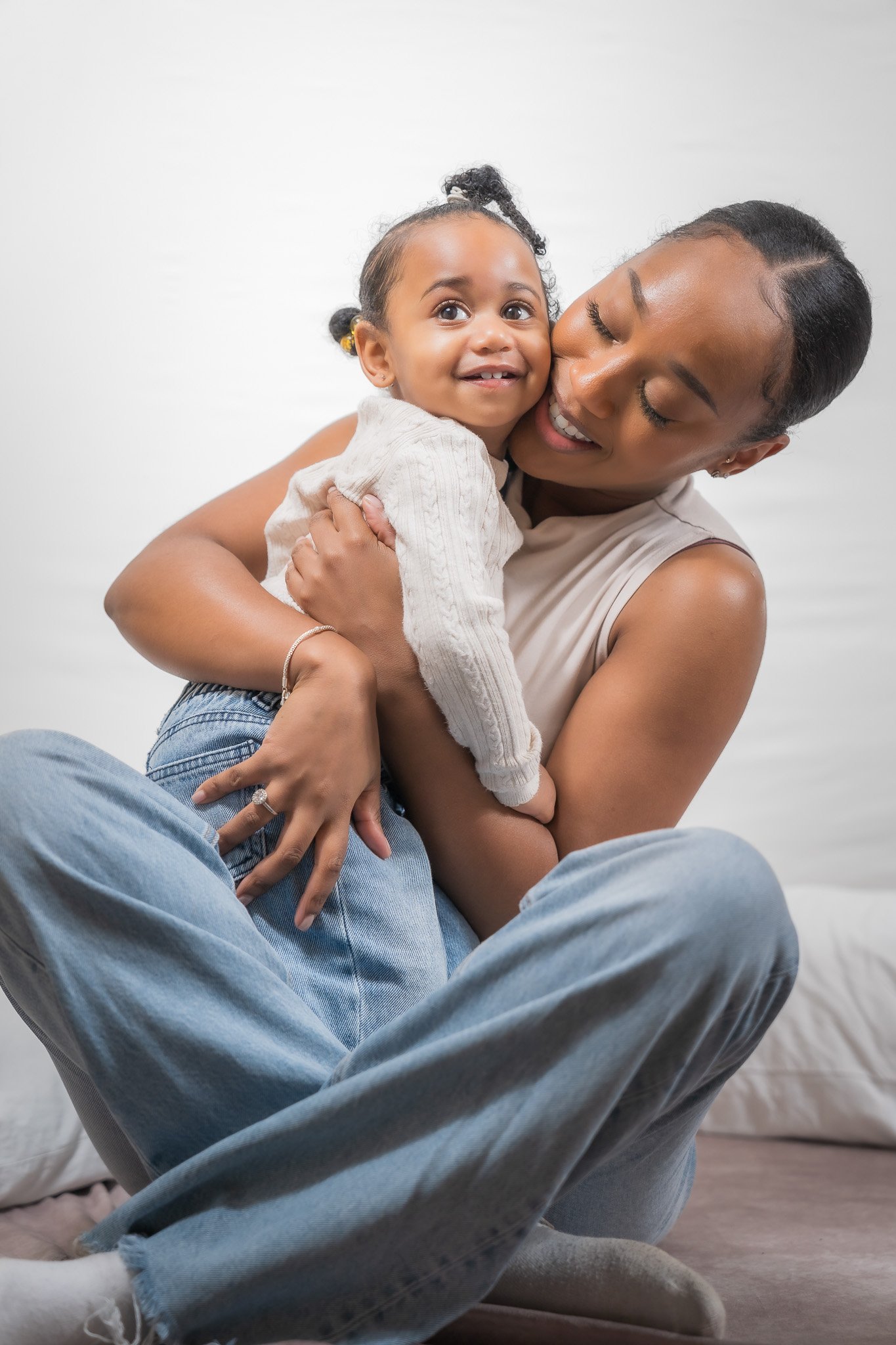 A woman with short hair holding and hugging a young girl with curly hair, smiling in a cozy indoor setting.